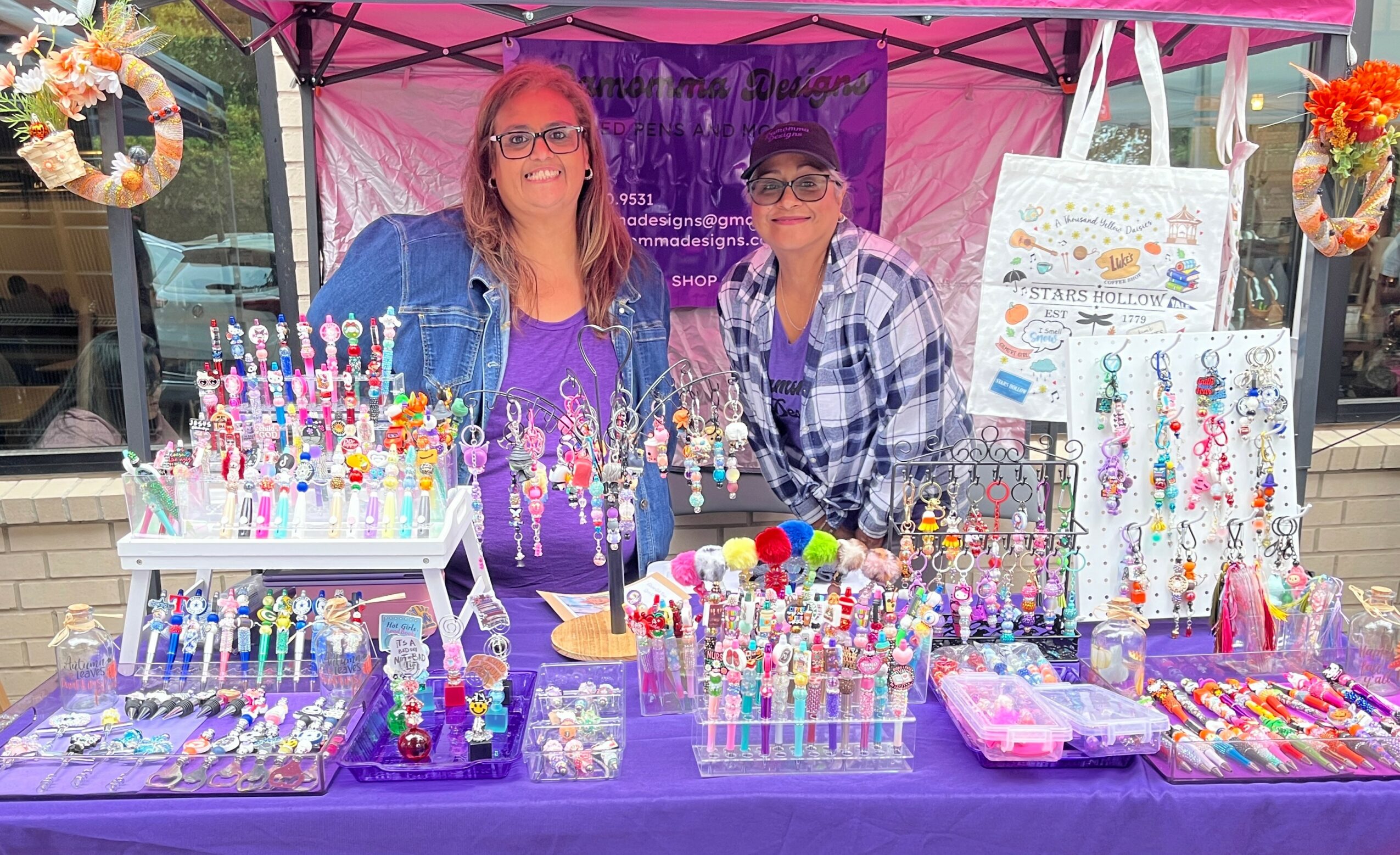 Two women stand behind a colorful jewelry display at an outdoor market stall, with jewelry and accessories on purple tablecloths.
