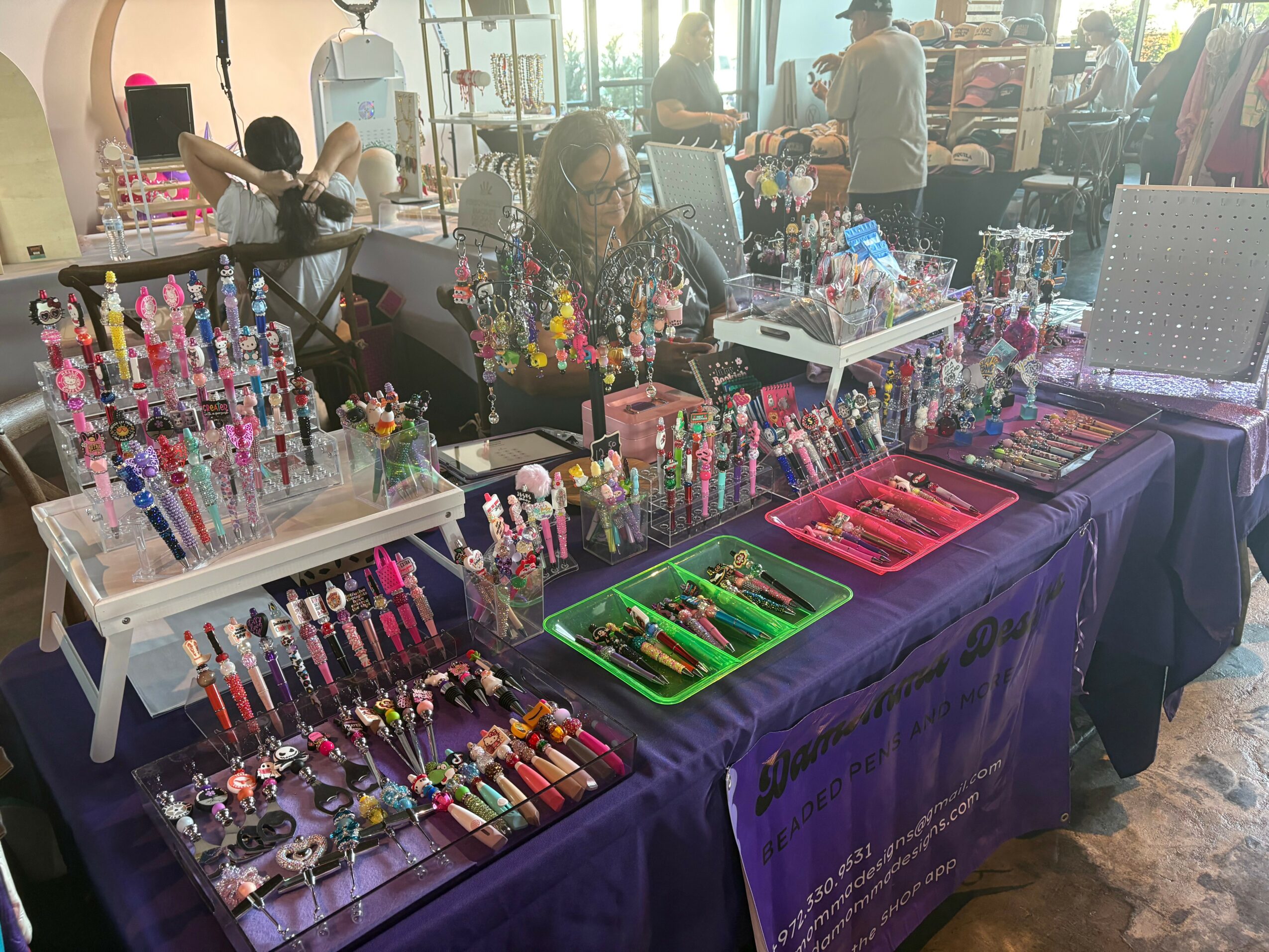 Market stall with colorful pens, watches, and accessories on tables covered with purple cloth, people browsing in background.