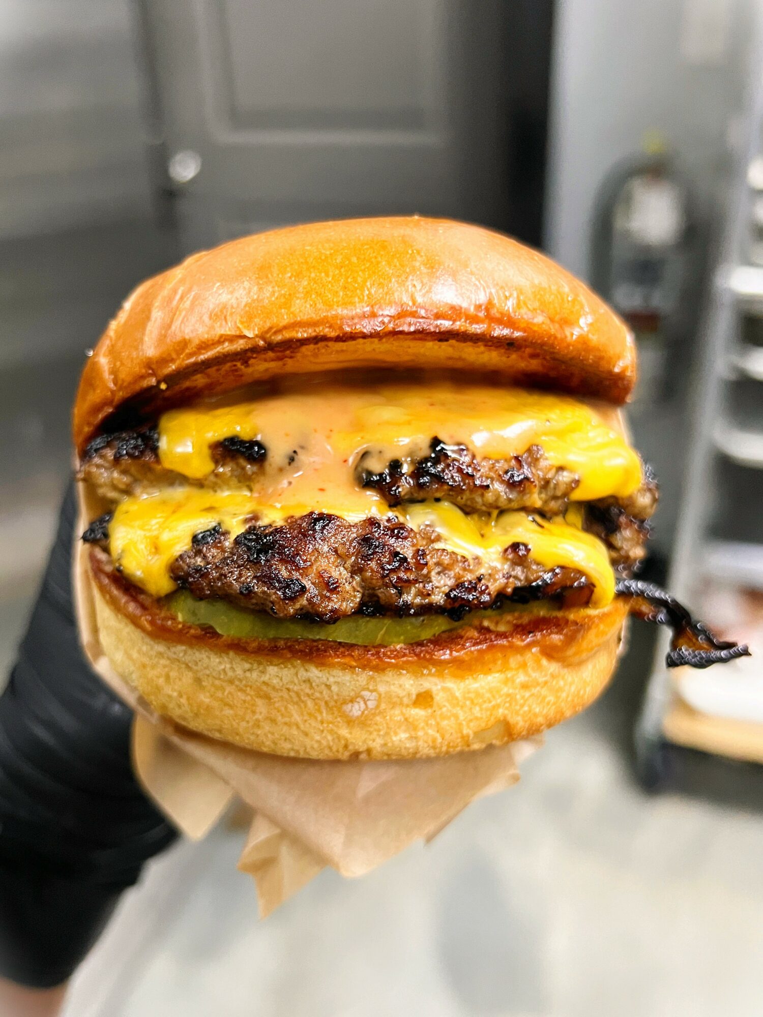 Close-up of a cheeseburger with melted cheese and beef patty in a bun, held by a hand in a kitchen.