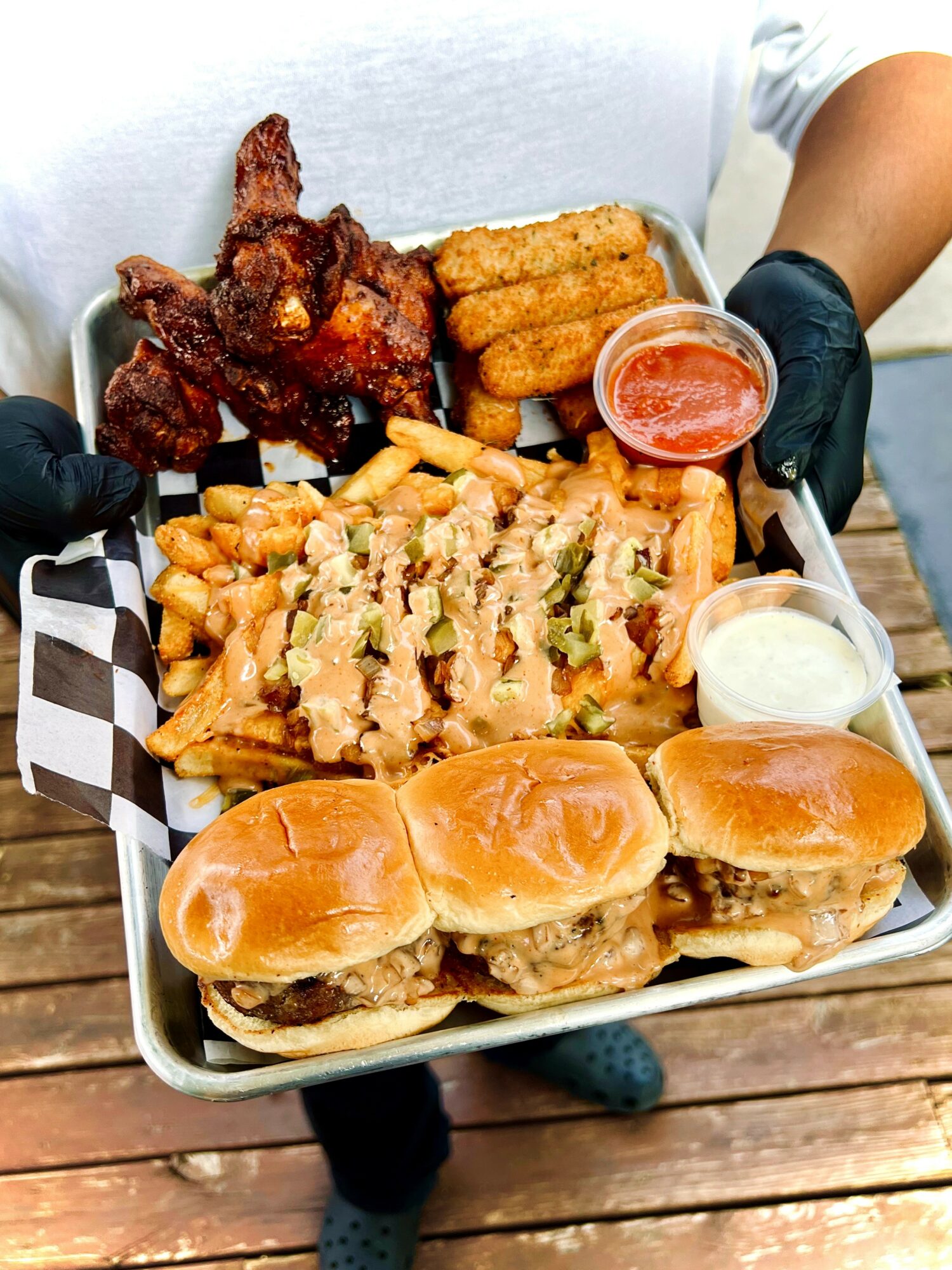 Person holding a tray with fried chicken, mozzarella sticks, fries, sliders, and dipping sauces, on a wooden surface.