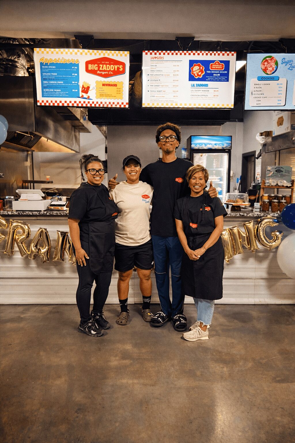 Four people standing in front of a restaurant counter with menu boards above, smiling, with a gold banner behind them.