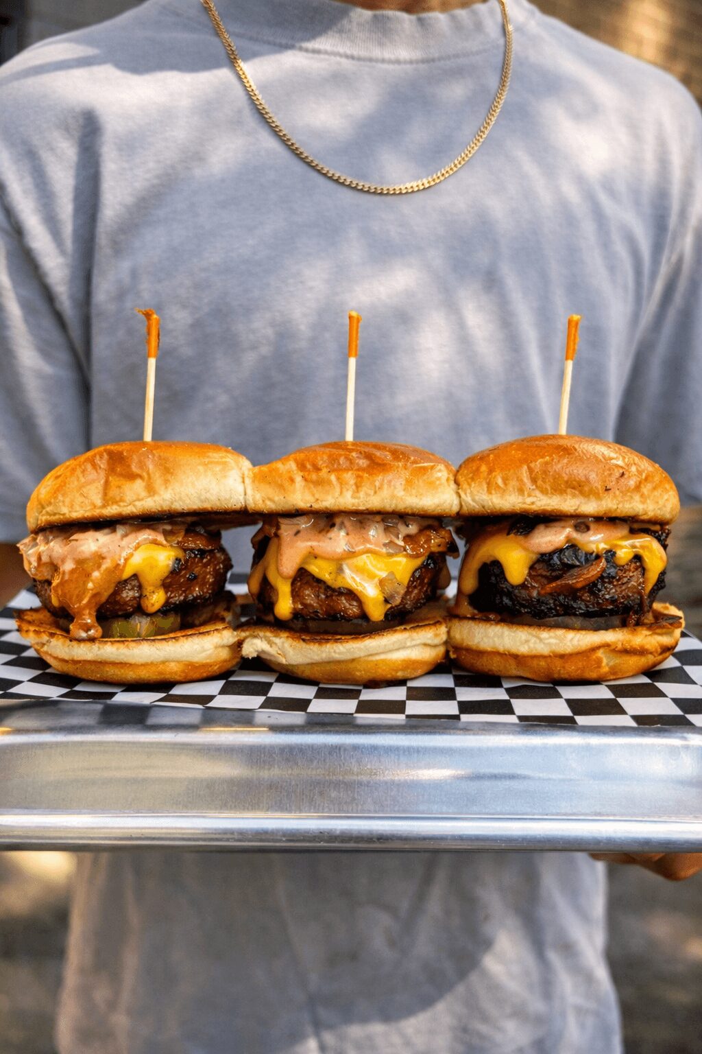 Three cheeseburgers with bacon and cheese on a checkered tray, person wearing a white shirt and gold chain in background.