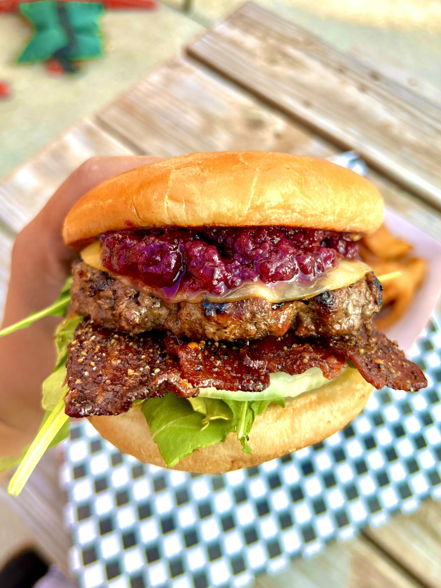 Hand holding a burger with beef patty, bacon, purple sauce, lettuce, and bun, outdoors on wooden table.