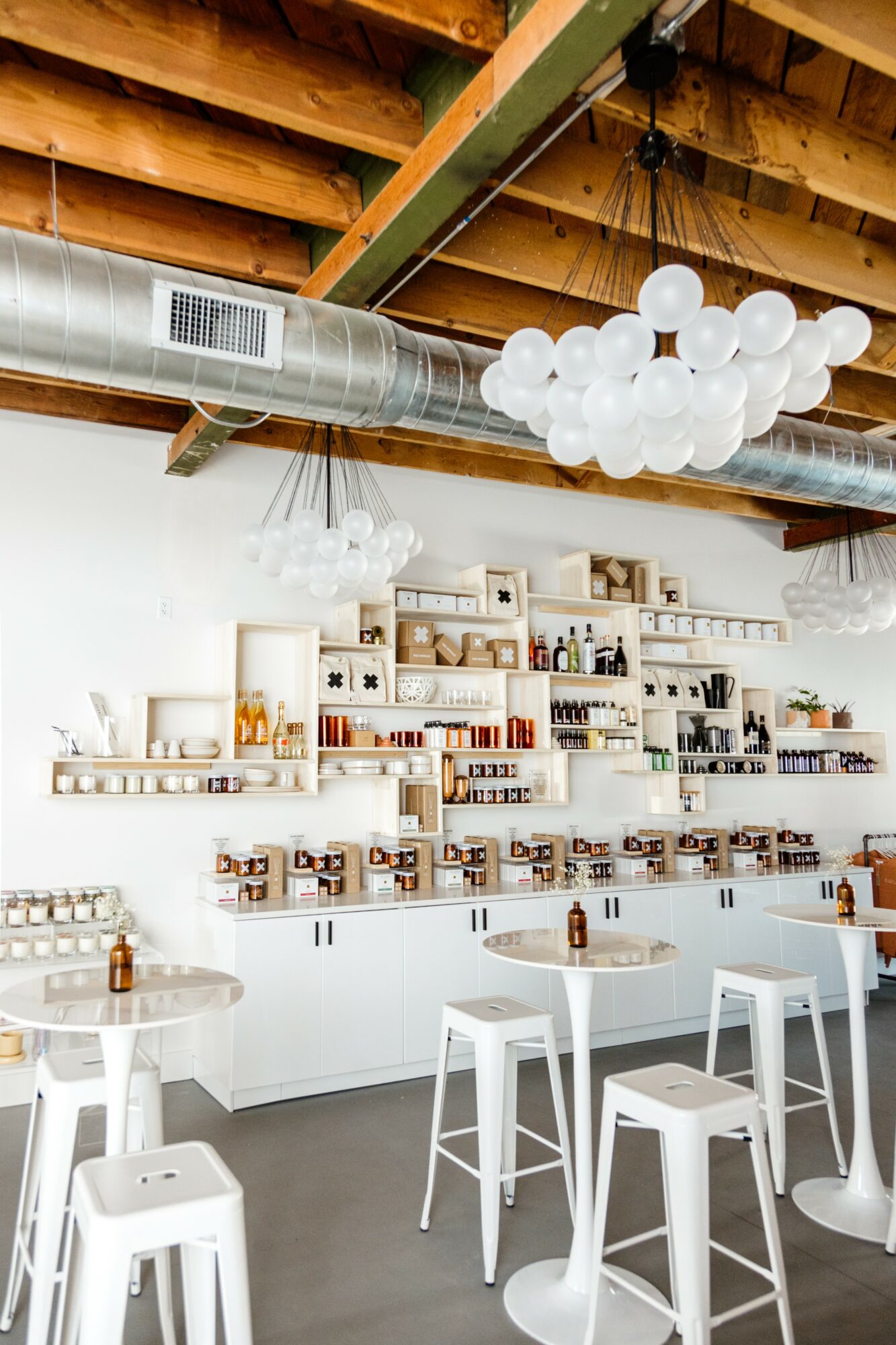 Interior of a modern cafe with white tables, chairs, and shelves displaying various products, ceiling with wooden beams and cloud-shaped light fixtures.