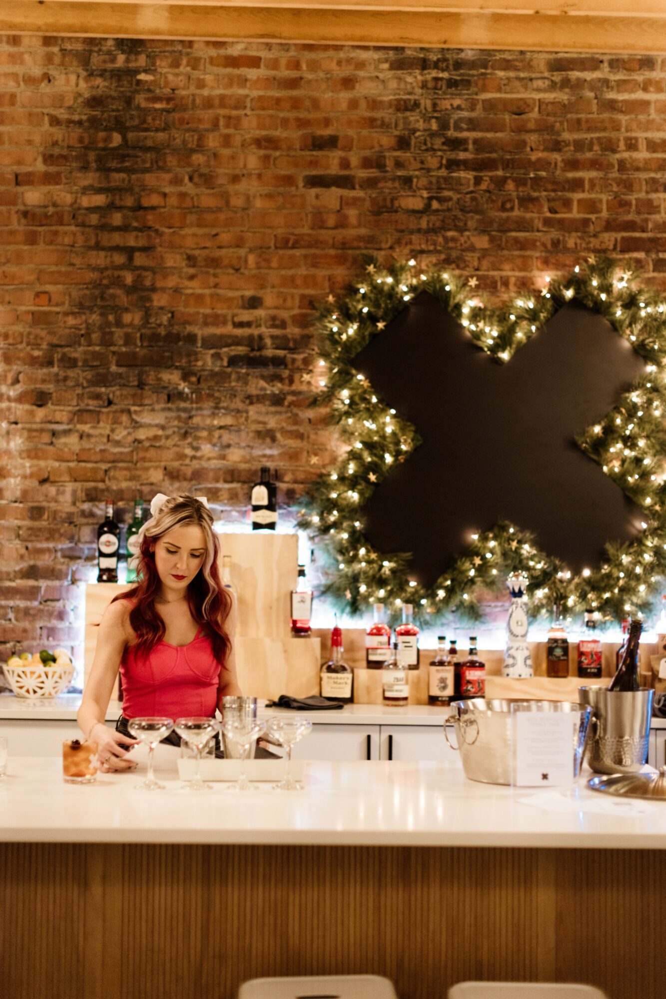 Young woman in red top standing behind a kitchen counter with a brick wall and decorated chalkboard in the background.