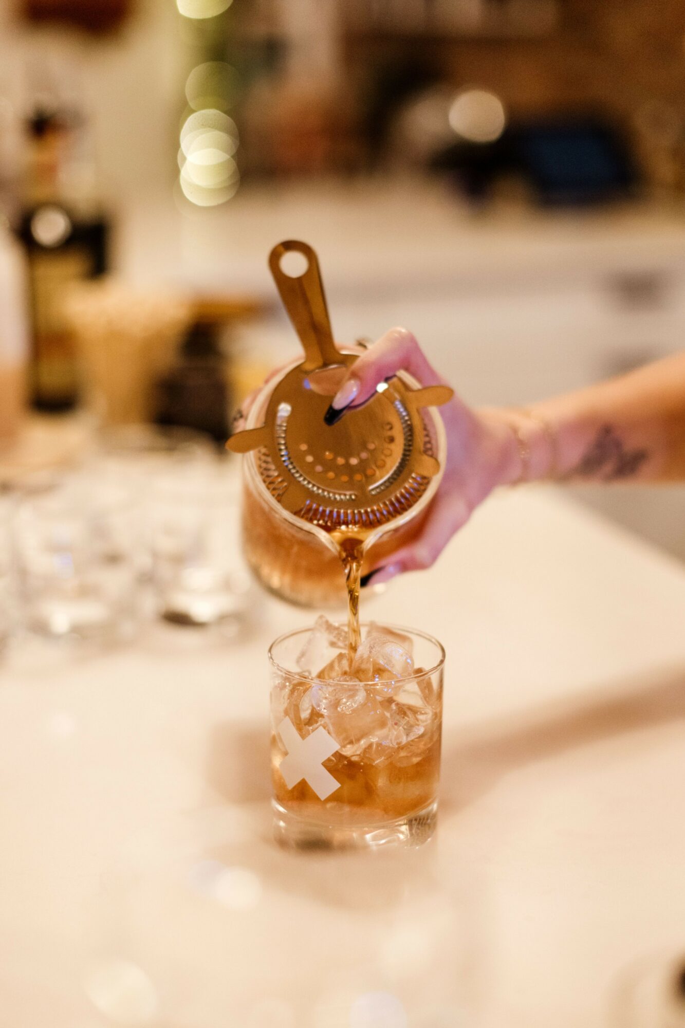 Person pouring a drink with a pink cocktail umbrella into a glass filled with ice, in a cozy bar setting.