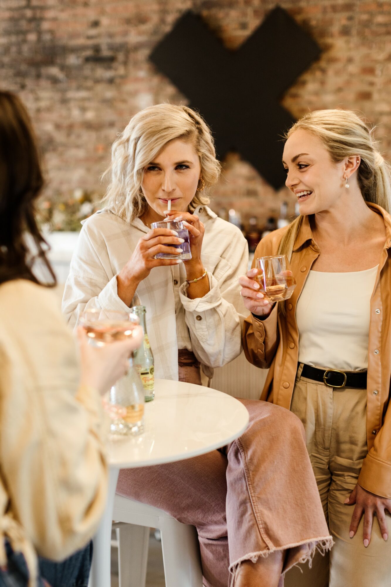Three women socializing at a bar, one drinking from a glass, smiling, in a cozy brick-walled setting.