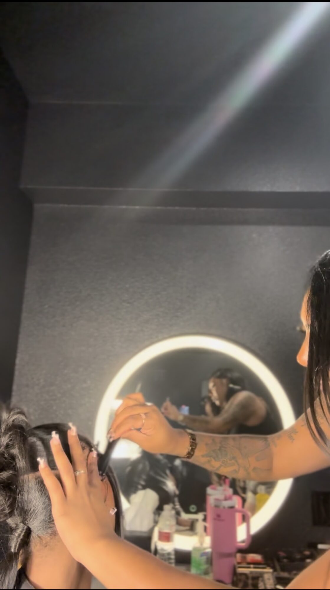 Person styling a child's hair in front of a round illuminated mirror, with hair tools and bottles on the counter.