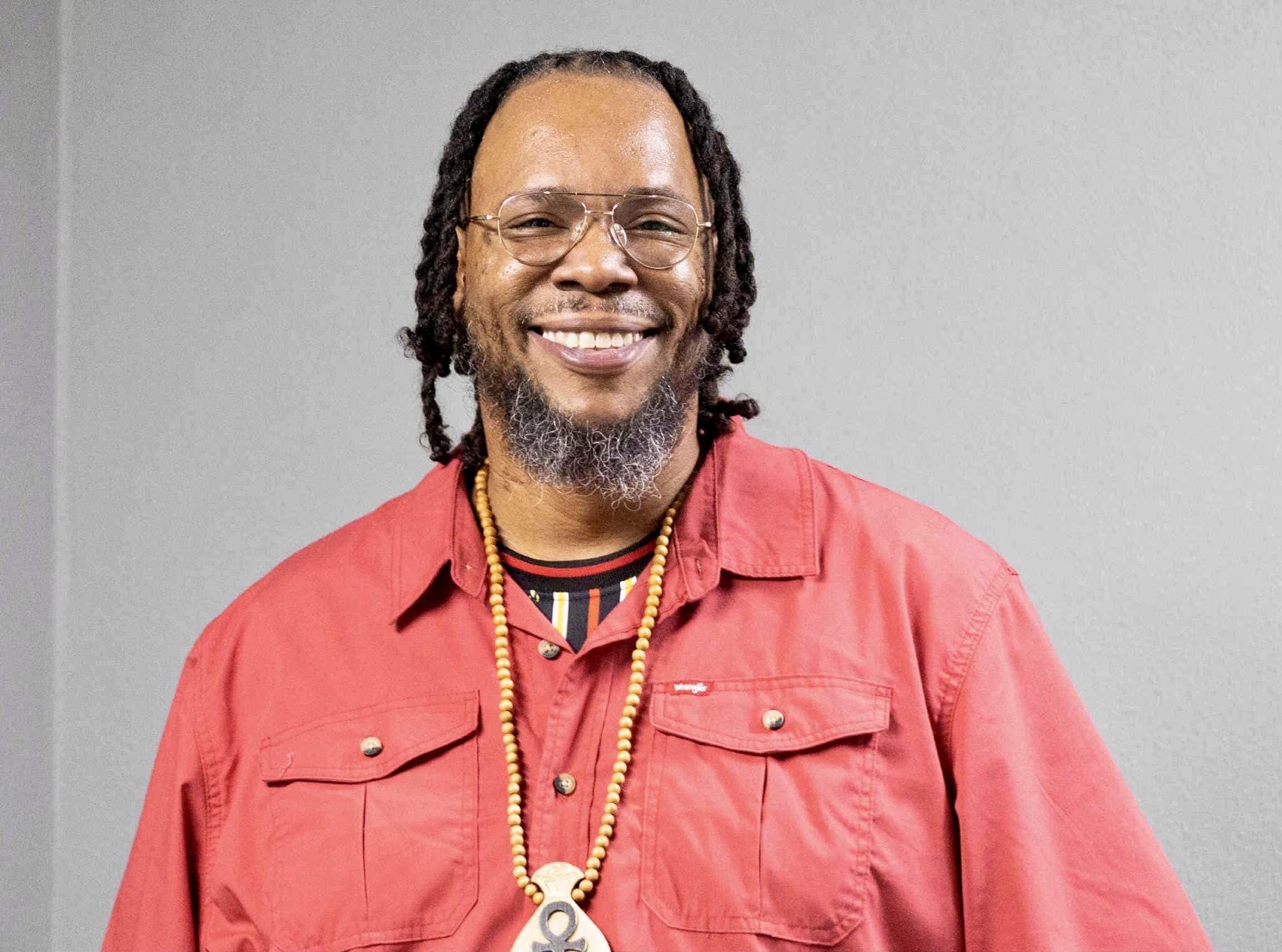 Smiling man with glasses, dreadlocks, wearing a red shirt and gold necklace, standing against a gray background.