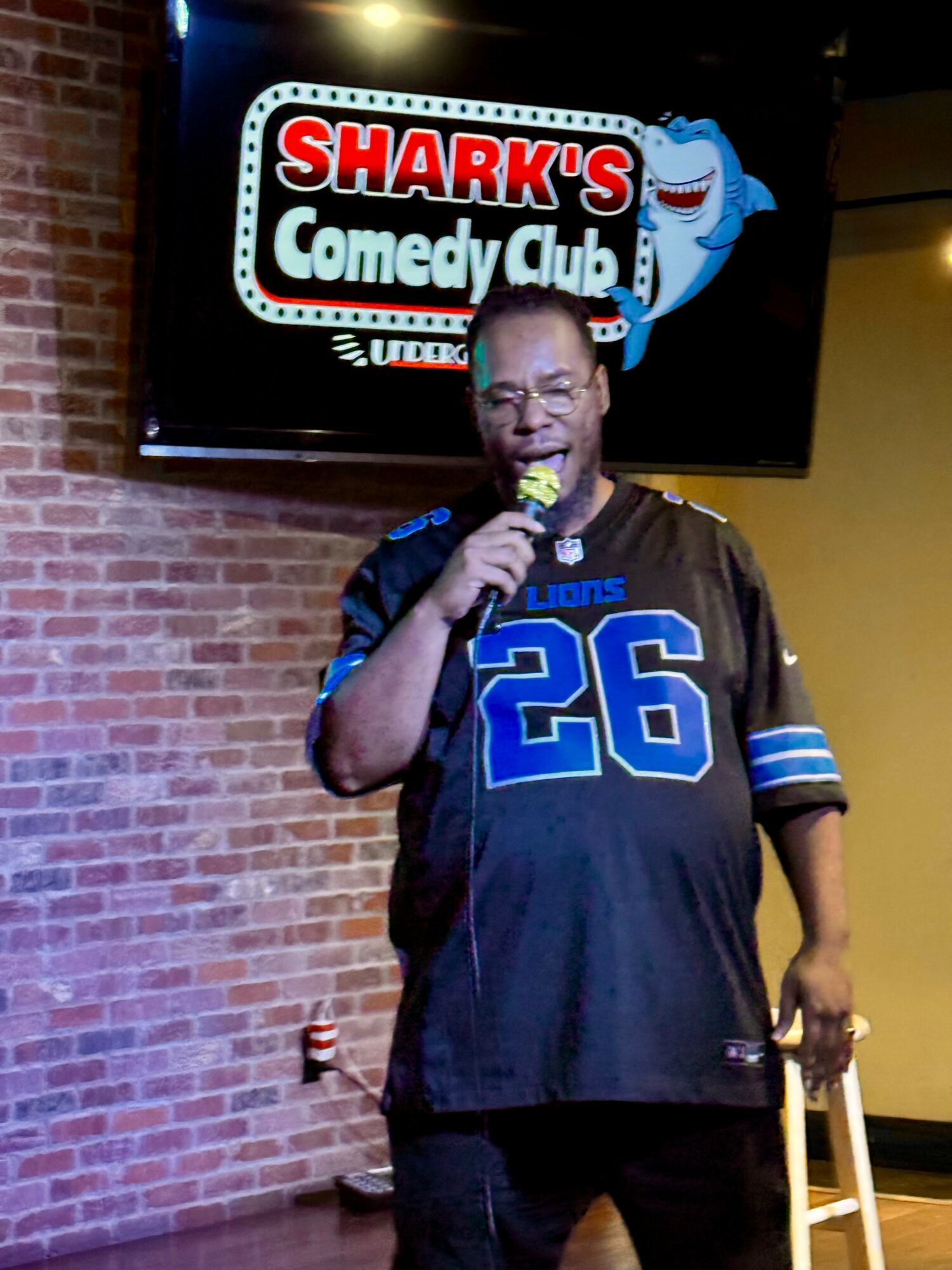 Man holding microphone on stage with Shark's Comedy Club sign behind him, brick wall background, wearing a black sports jersey with blue number 26