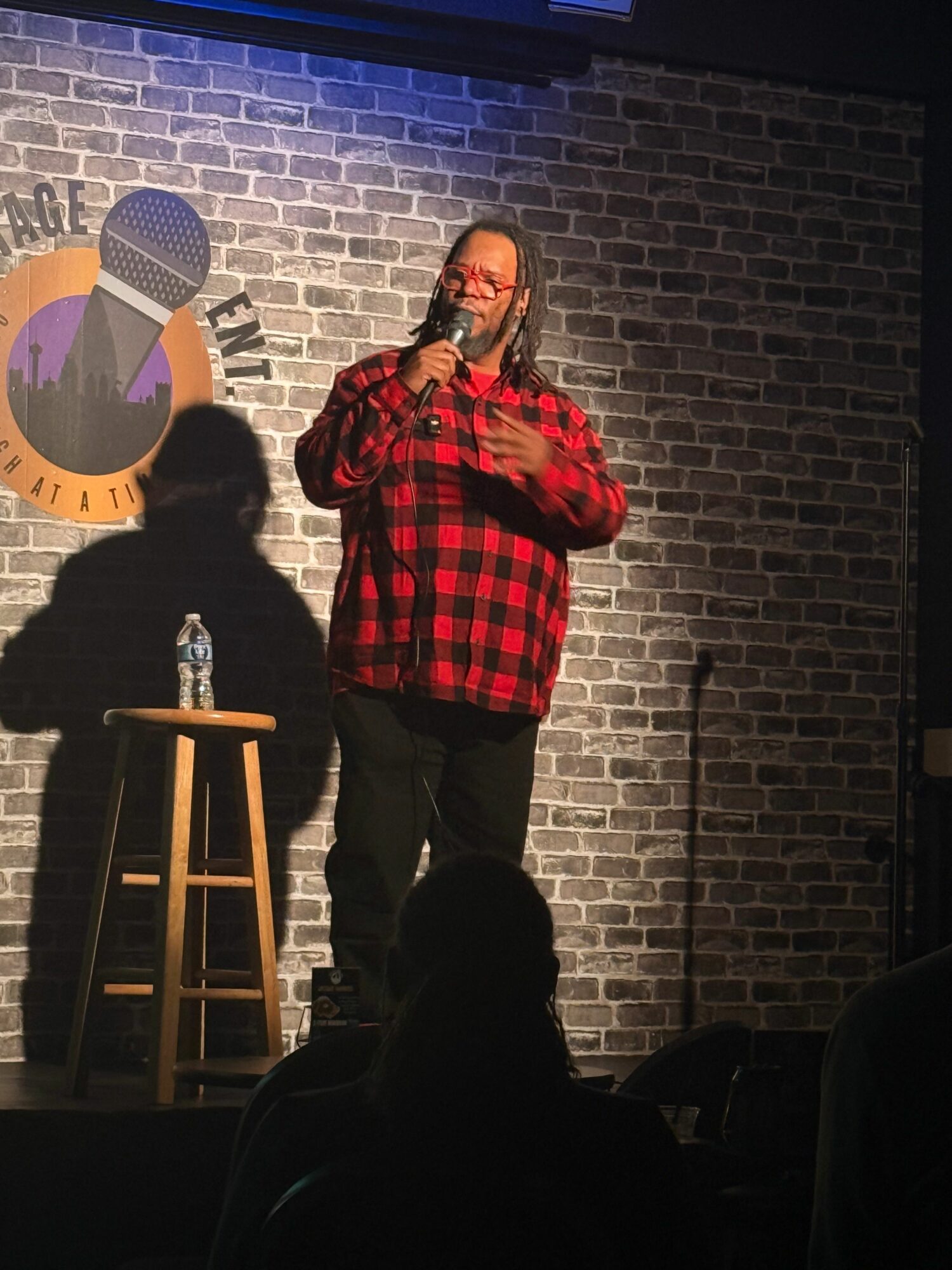 Person in red plaid shirt standing on stage with brick wall background, holding a microphone, with a stool nearby.