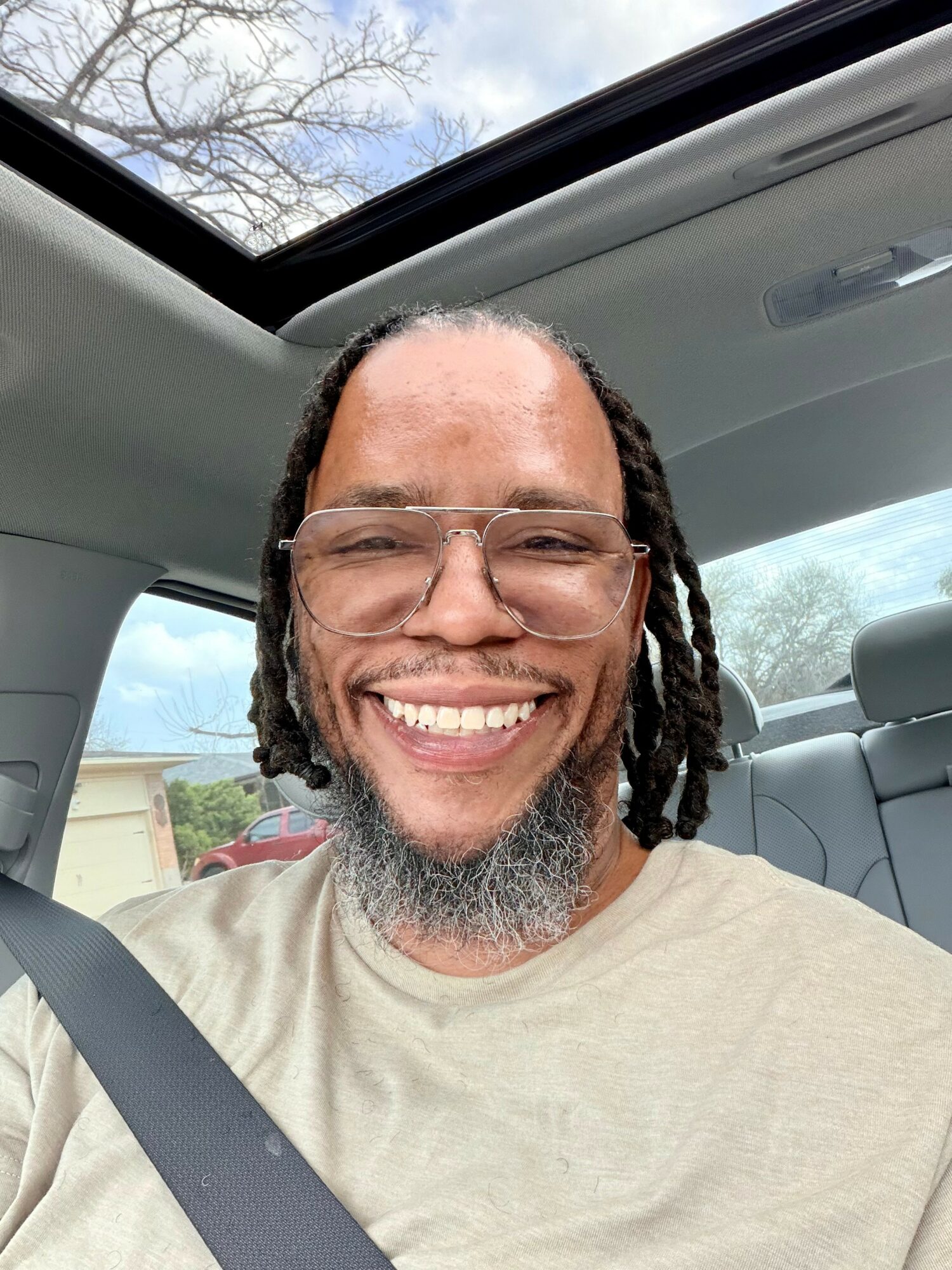 Smiling man with glasses and dreadlocks inside a car, with a sunroof showing trees and sky.