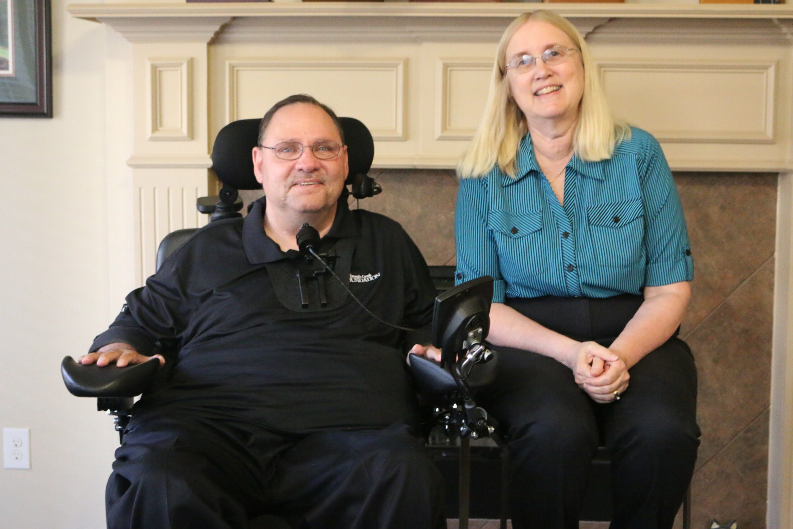 A man in a wheelchair and a woman sit together indoors, smiling, in front of a fireplace mantel.