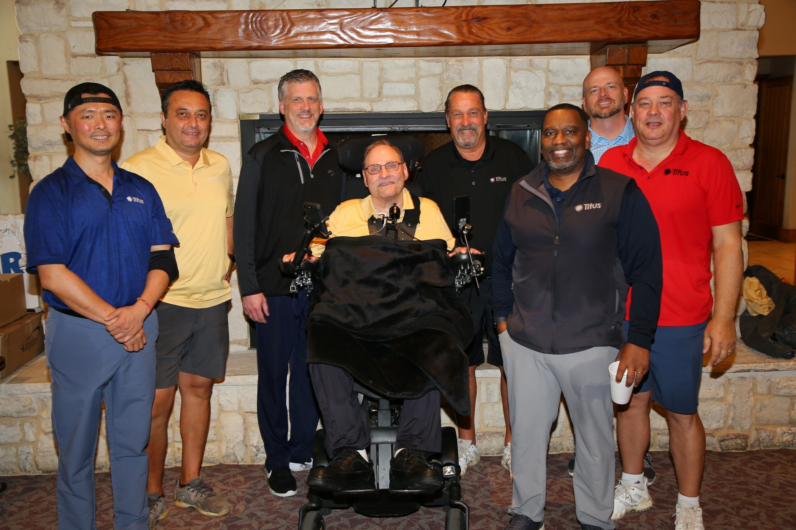 Group of eight people standing in front of a fireplace, smiling, with one person in a wheelchair in the center.