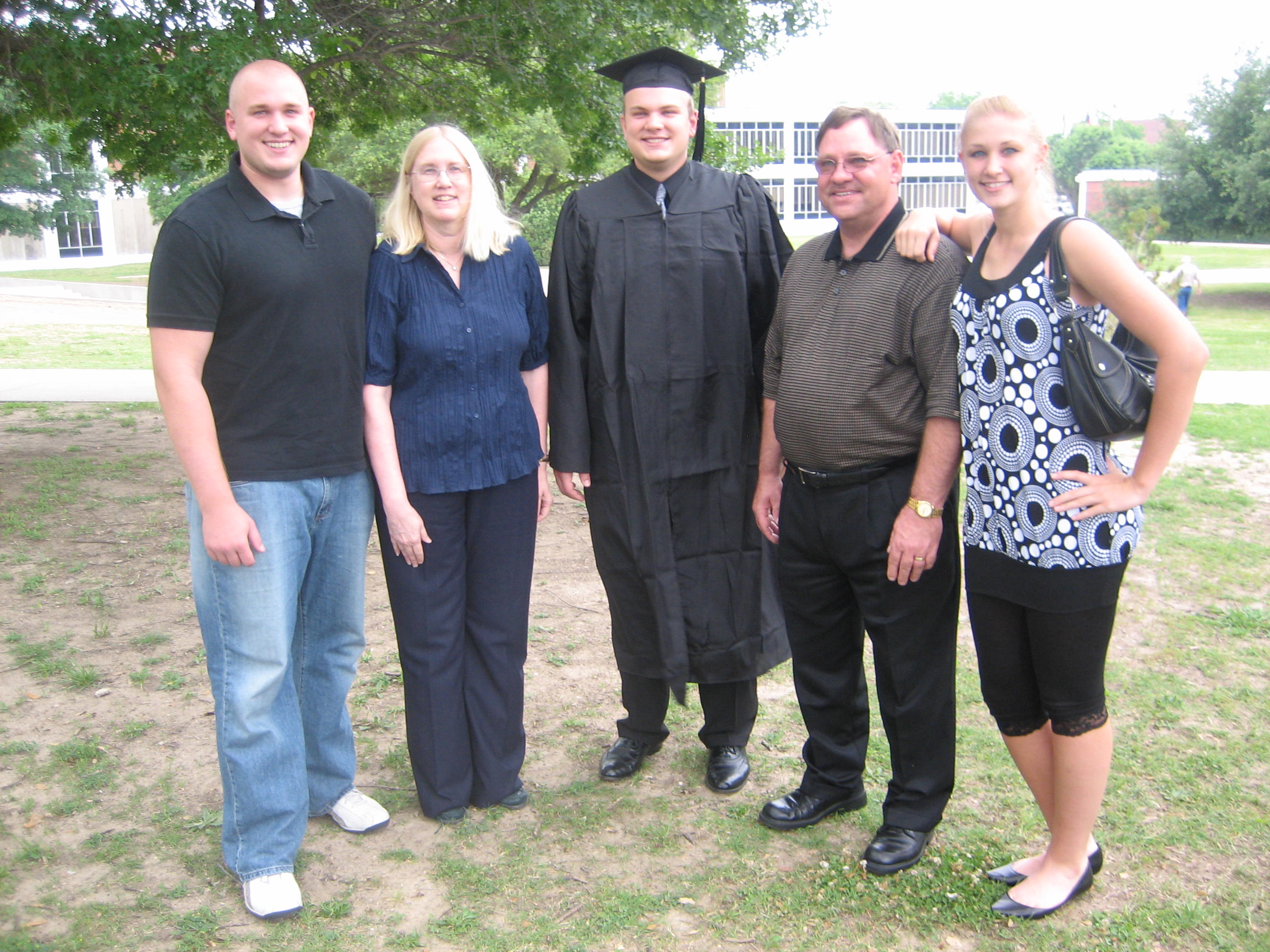 Group of five people outdoors, one in graduation gown and cap, others in casual clothing, standing on grass with trees.