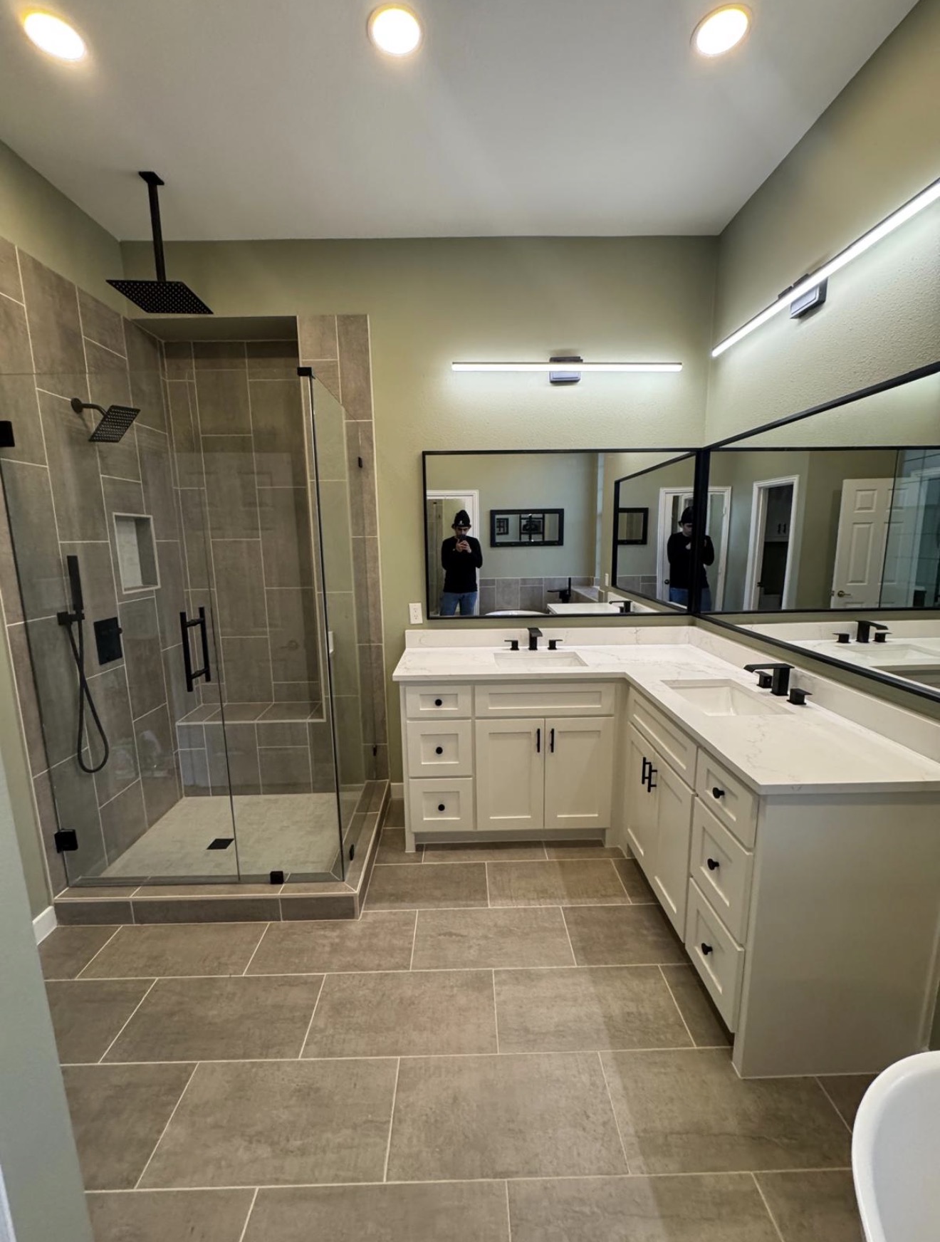 Bathroom with shower, double sink vanity, large mirror, and tiled floor, seen from an angle. Two people are reflected in the mirror.