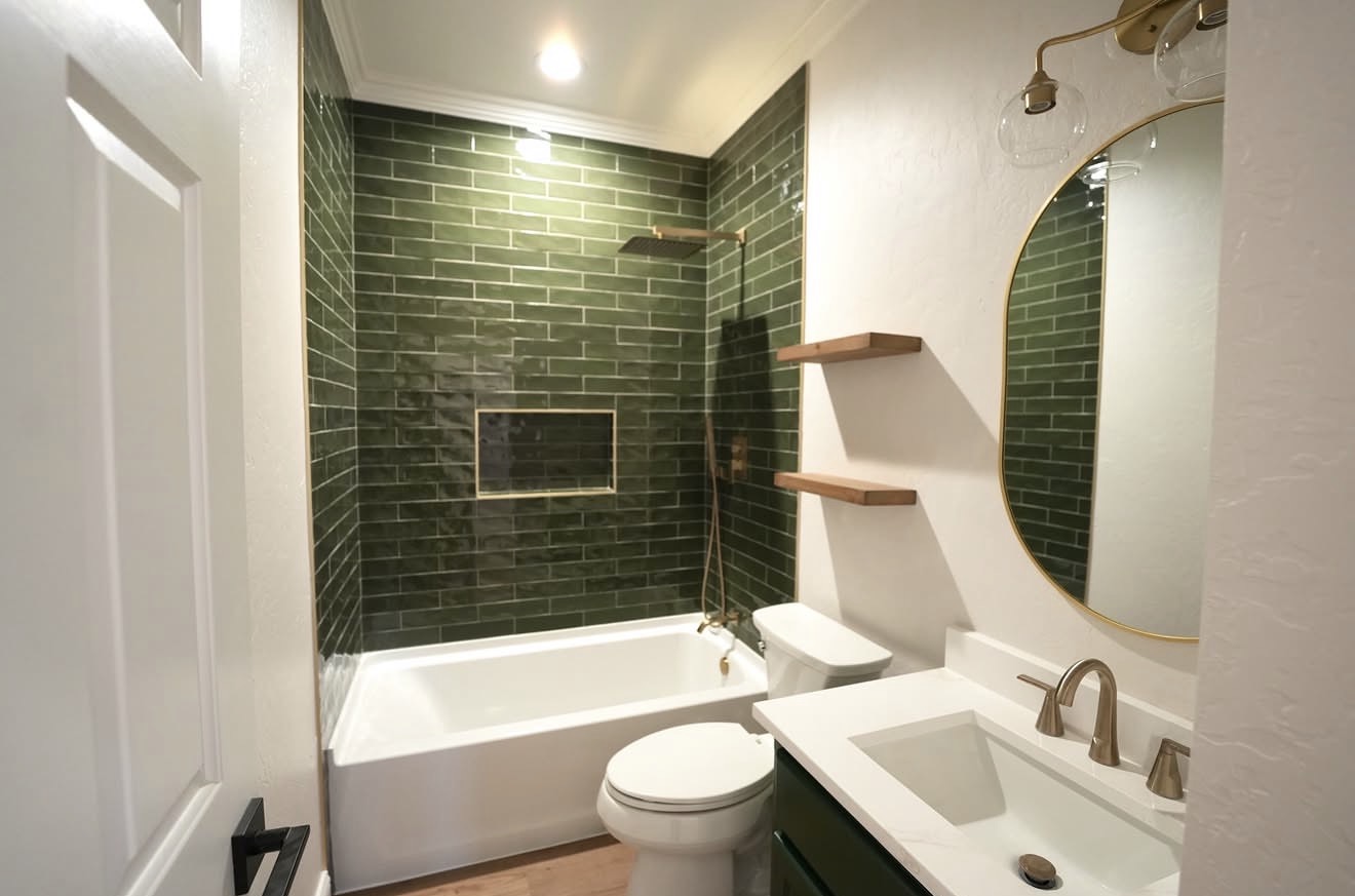 Bathroom with green tiled shower area, white bathtub, toilet, and a white sink with a round mirror above.