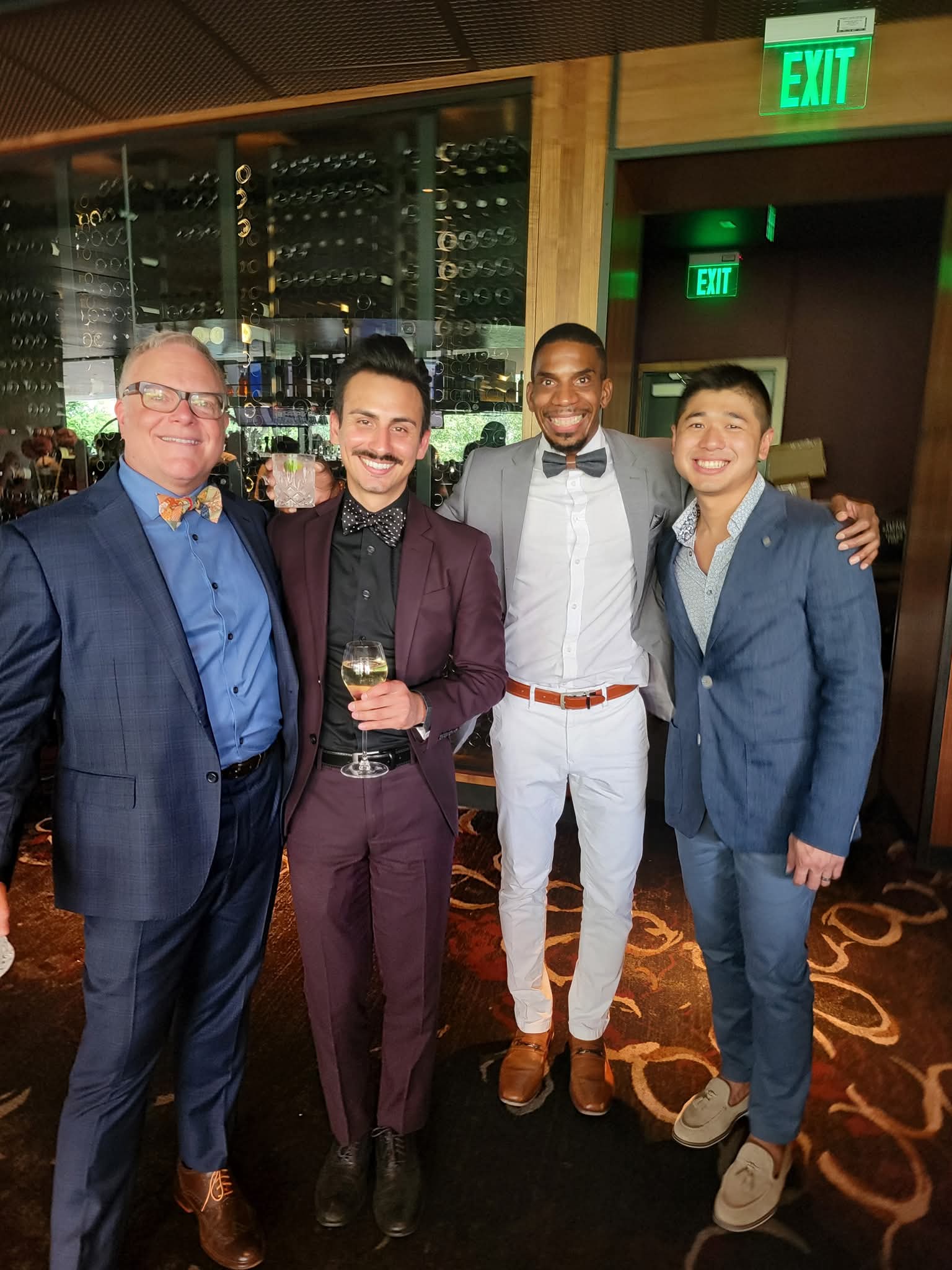Four men in formal attire standing together in a bar or lounge, smiling, with wine bottles and glasses in the background.