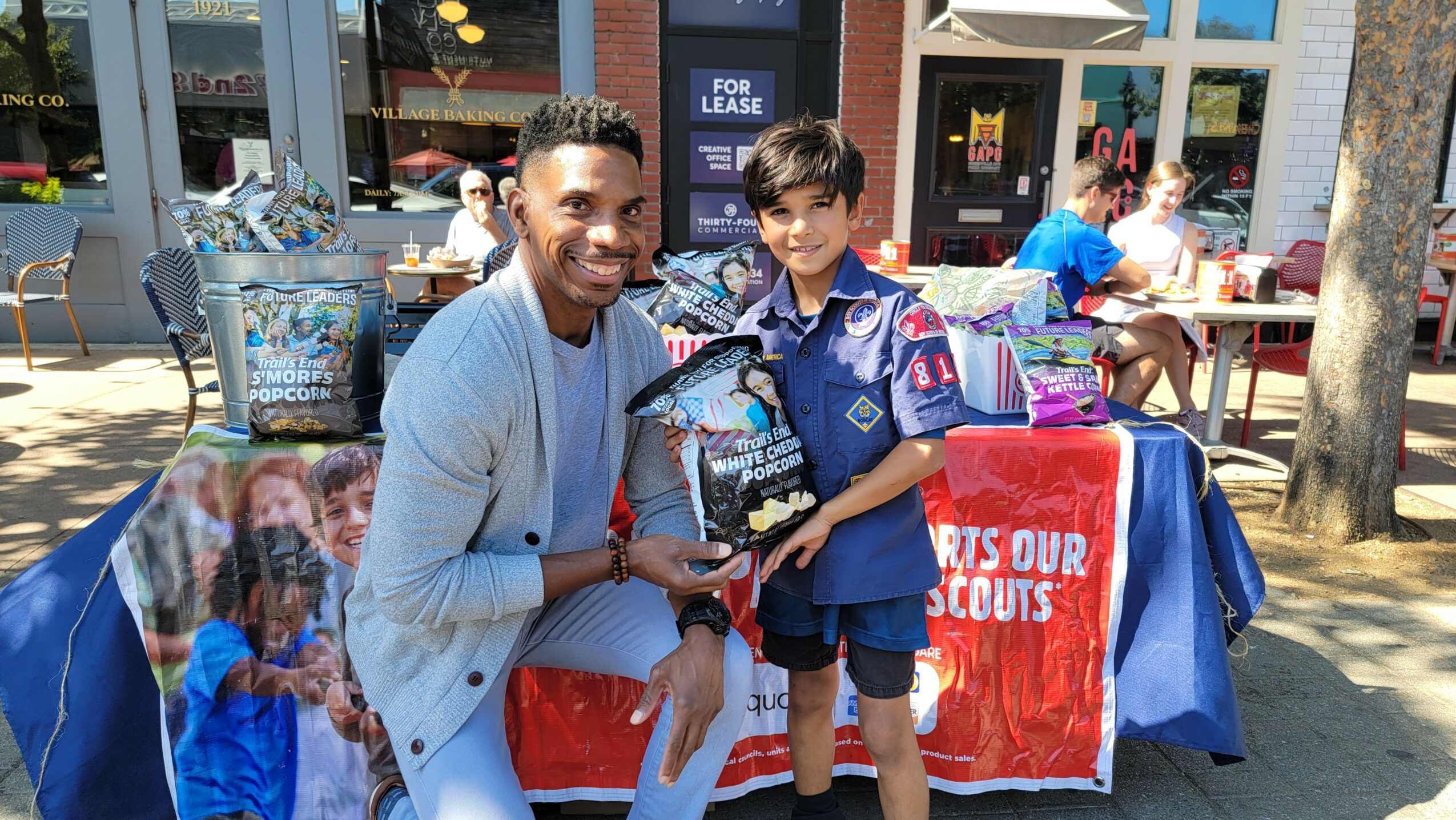 Two people, a man and a boy, standing outdoors near a table with a red tablecloth, holding a small gift bag.