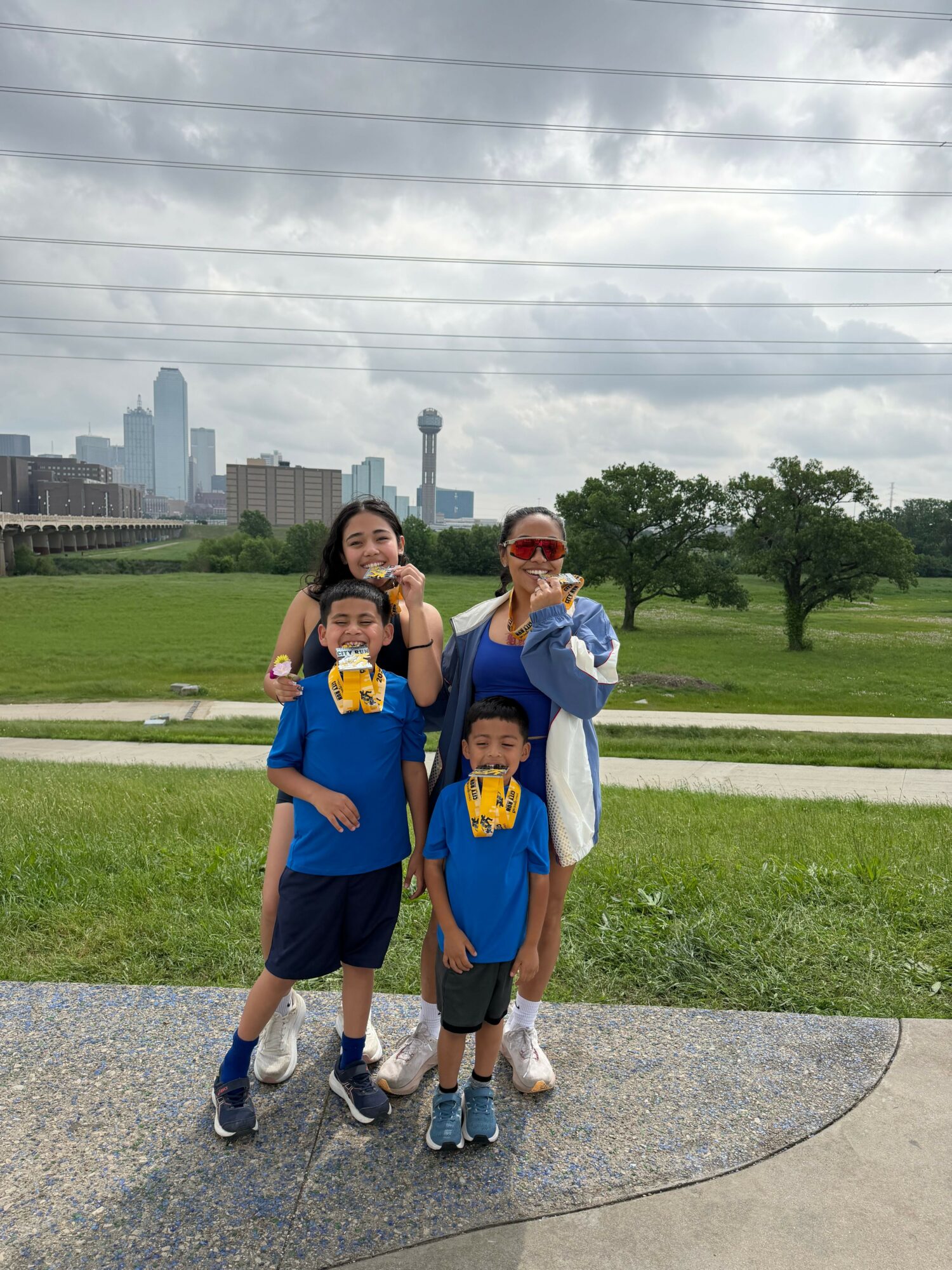 Two children and an adult standing outdoors on a sidewalk, with a grassy area and trees in the background.
