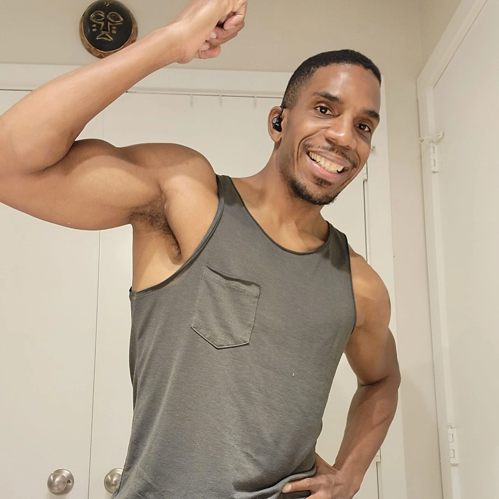 Man smiling, flexing arm, wearing sleeveless shirt, in a room with white walls and a decorative wall hanging.