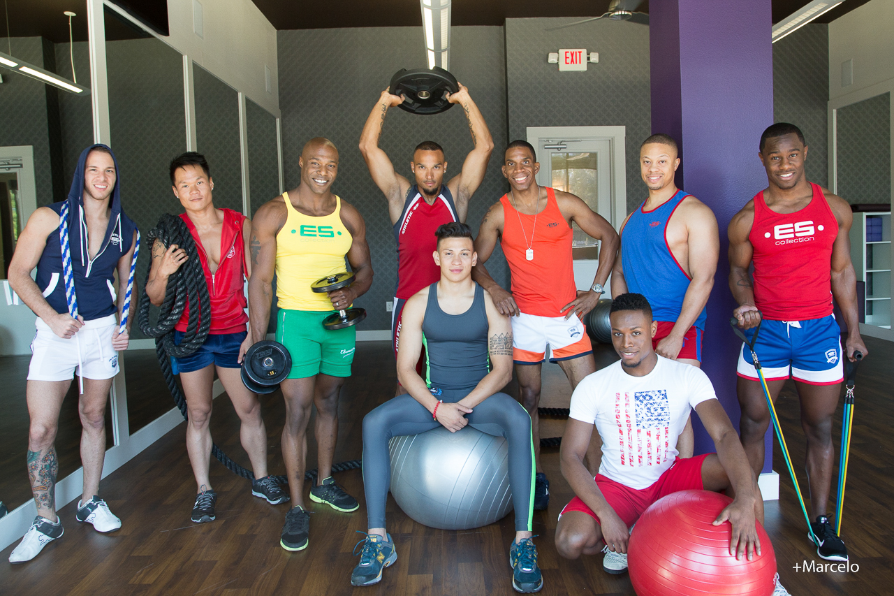 Group of eight young people in gym, some holding weights, one sitting on exercise ball, others standing with fitness equipment.