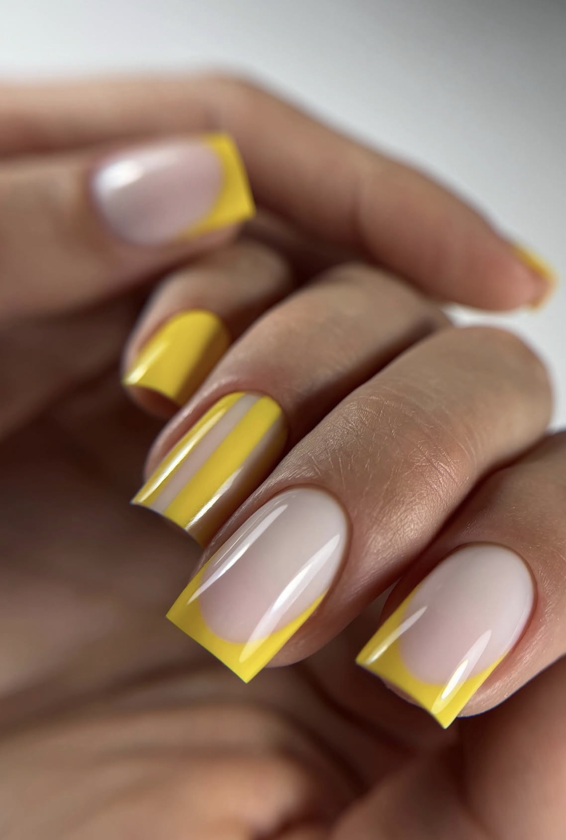 Close-up of a hand with nails painted yellow and clear, with yellow tips, against a neutral background.