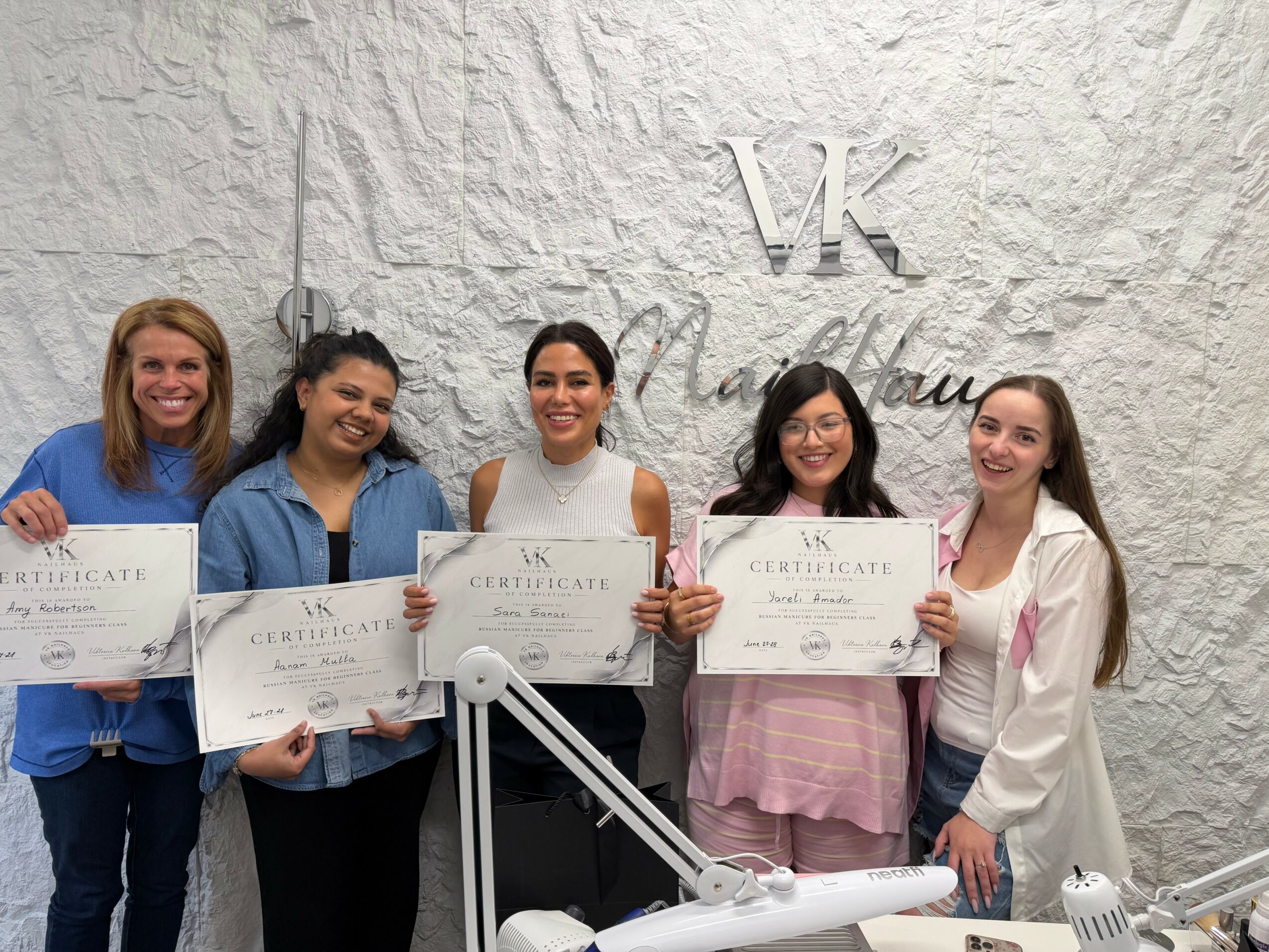 Five women holding certificates and smiling in front of a textured wall with a logo. They are standing close together.