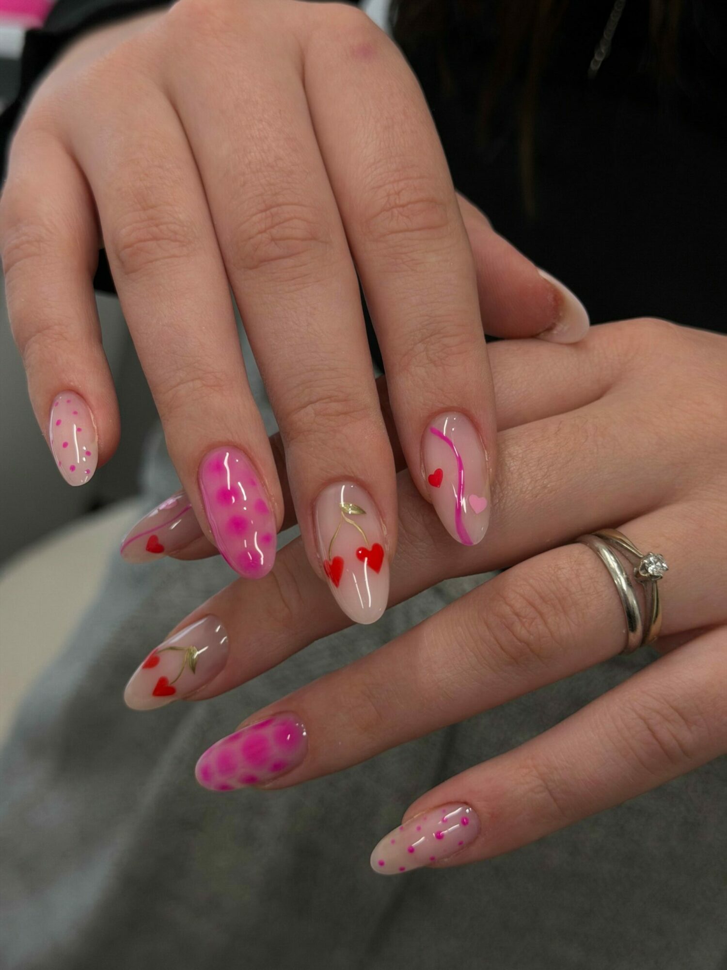 Two hands with decorated nails, some with pink and red heart designs, and rings on fingers.