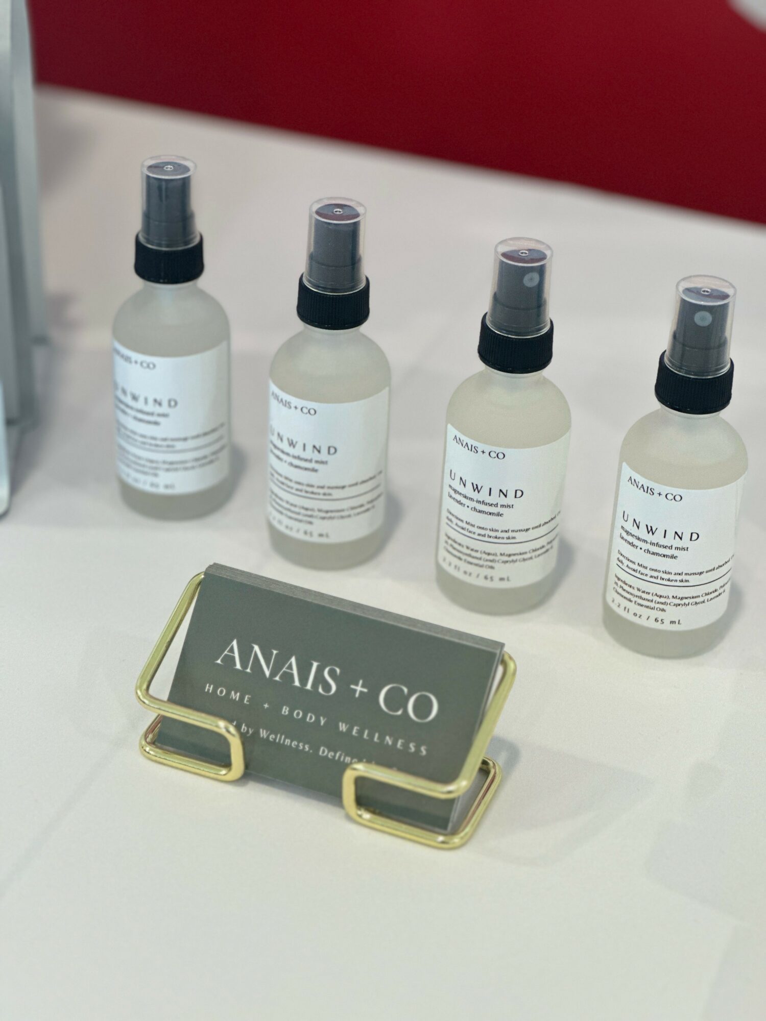 Four spray bottles with labels on a white surface, a black and gold sign reading 'ANAIS + CO' in front.