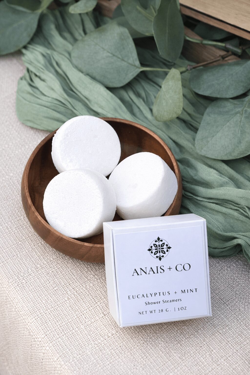 Three white bath bombs in a round wooden bowl, with eucalyptus leaves and a box of eucalyptus and mint soap nearby.
