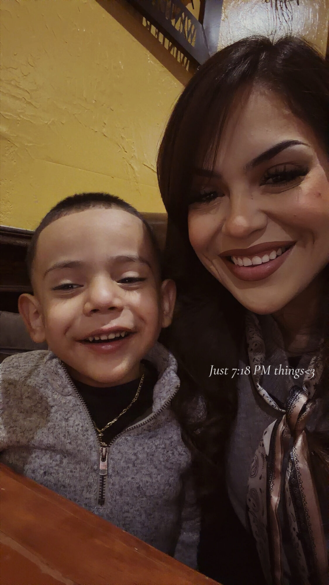 A smiling woman and young boy sit close together indoors, with a yellow wall in the background.