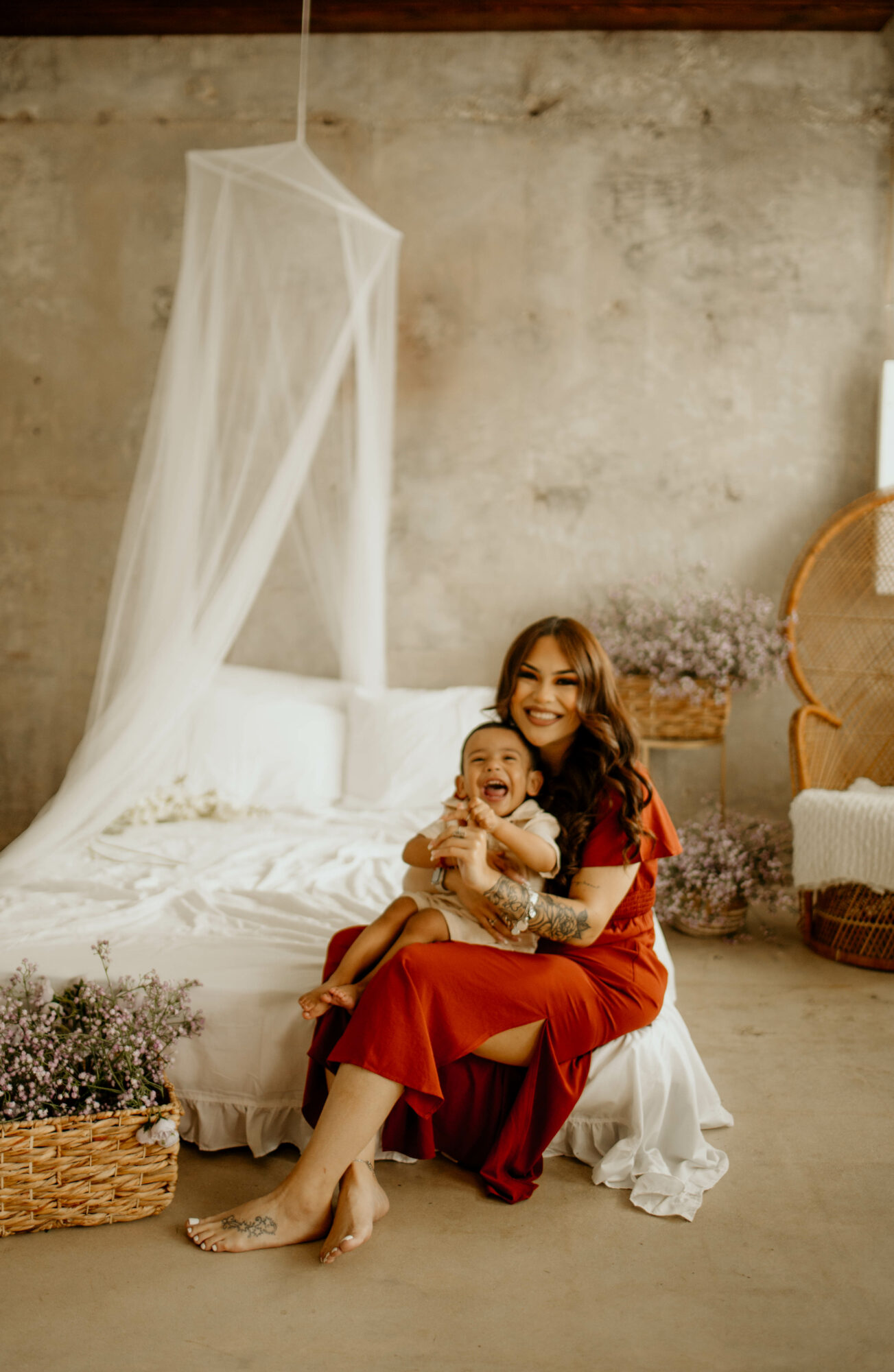 Woman sitting on bed holding a child, smiling, in a cozy room with a canopy bed and decorative plants.
