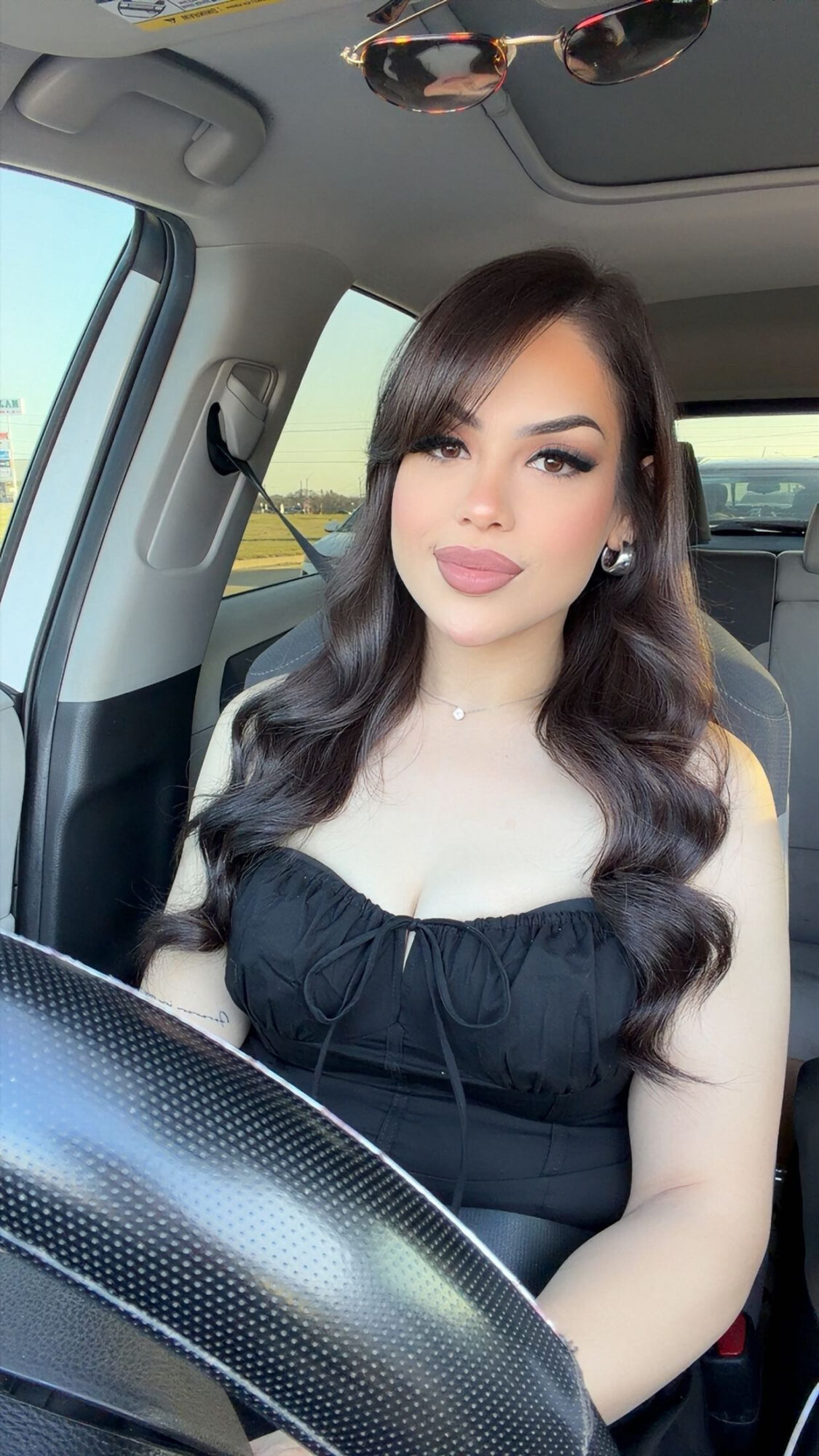 Young woman with long wavy dark hair and makeup, sitting in car, wearing a black top, looking at camera.