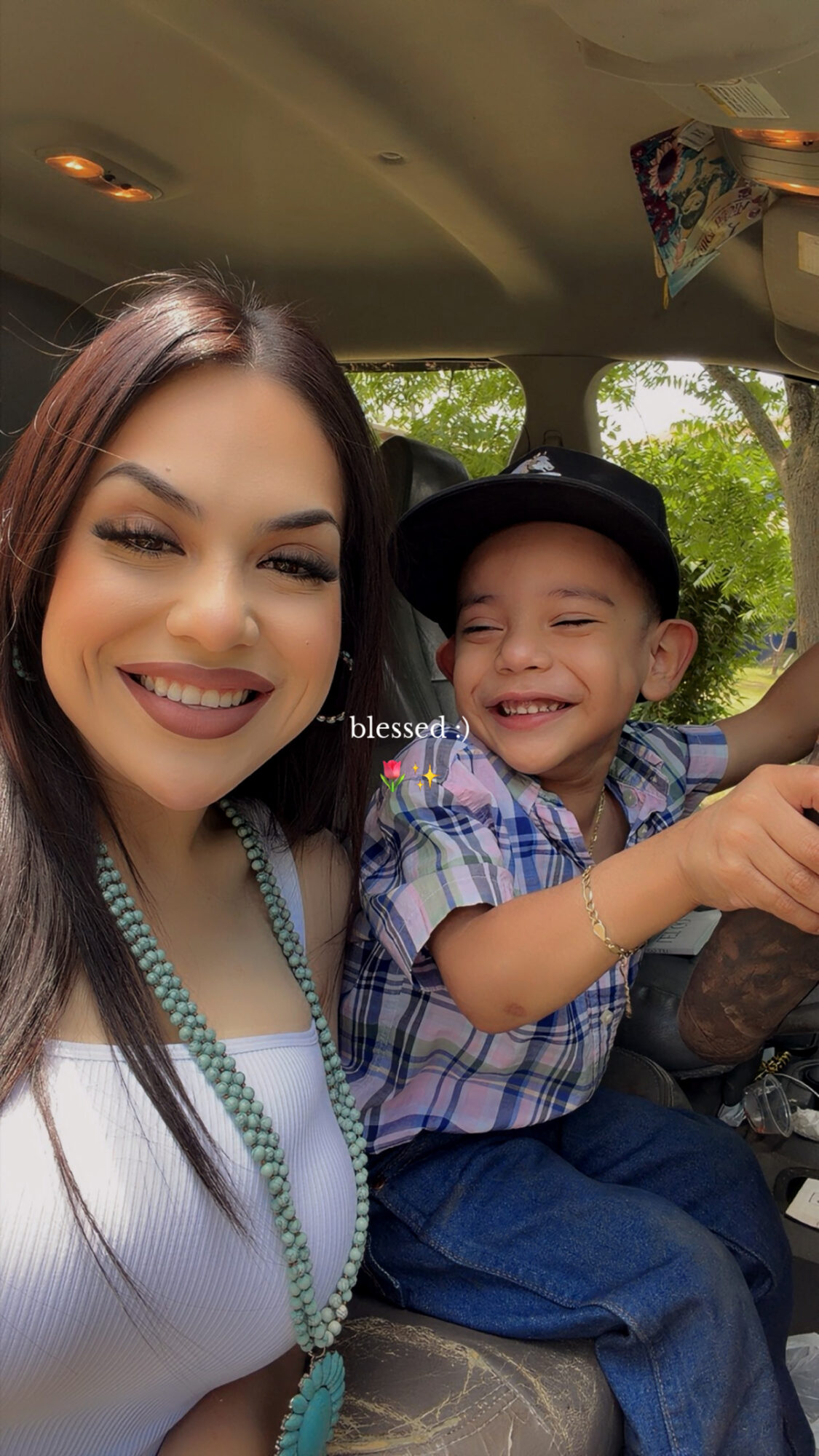 A woman and a young boy smiling inside a vehicle, with trees visible outside the window.