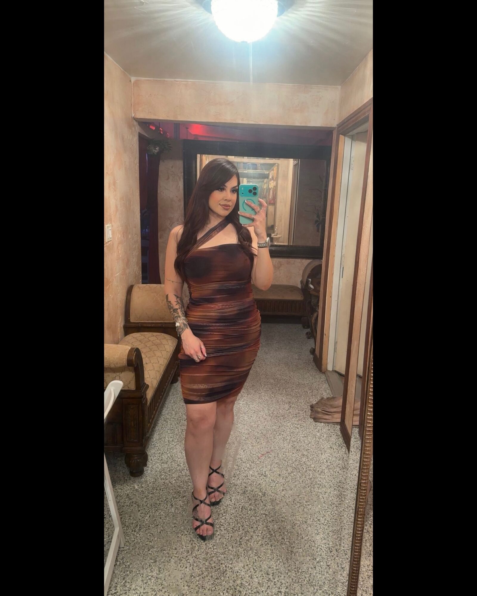 Woman taking a mirror selfie in a room with beige walls, wearing a fitted, multicolored dress and high heels.