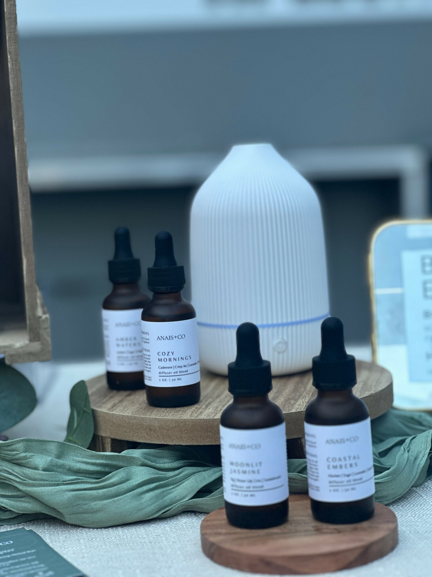A white humidifier and five small bottles with droppers on wooden platforms, surrounded by green fabric and a blurred background.