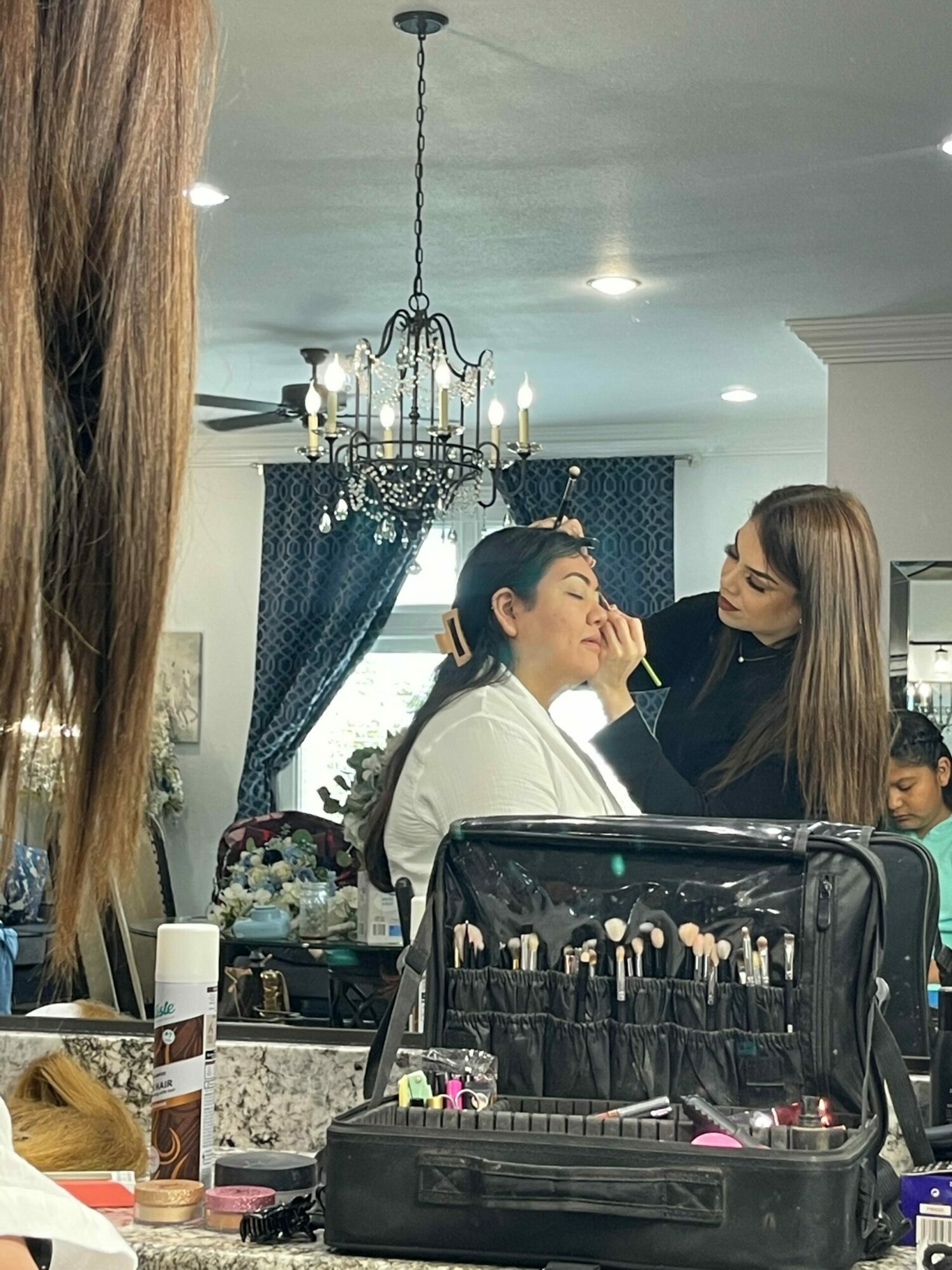 Two women are in a room with makeup and beauty products on a table, engaging in conversation. A chandelier hangs from the ceiling.