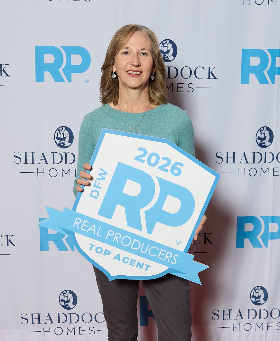 Woman holding a sign with '2026' and 'RP' at an event, smiling, standing in front of a backdrop with logos.