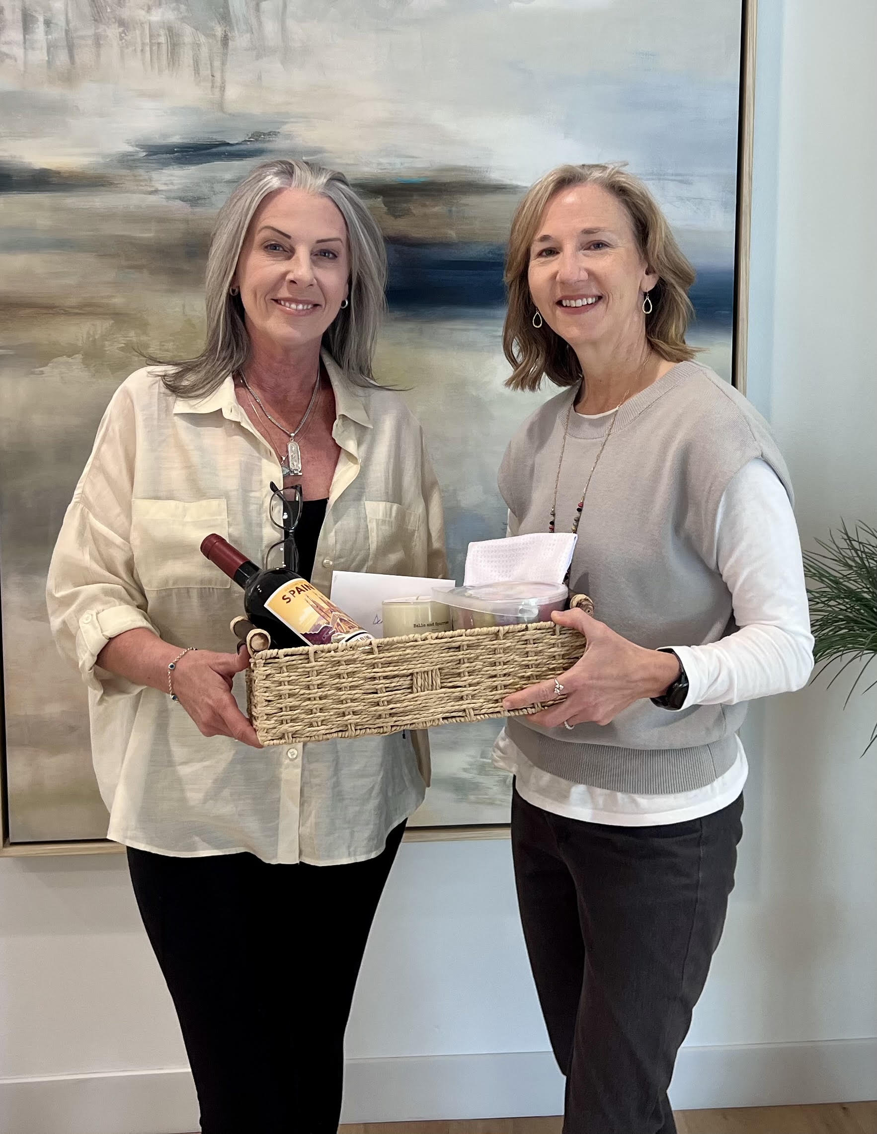 Two women smiling, holding a basket with a bottle of wine and a box, standing in front of a painting.