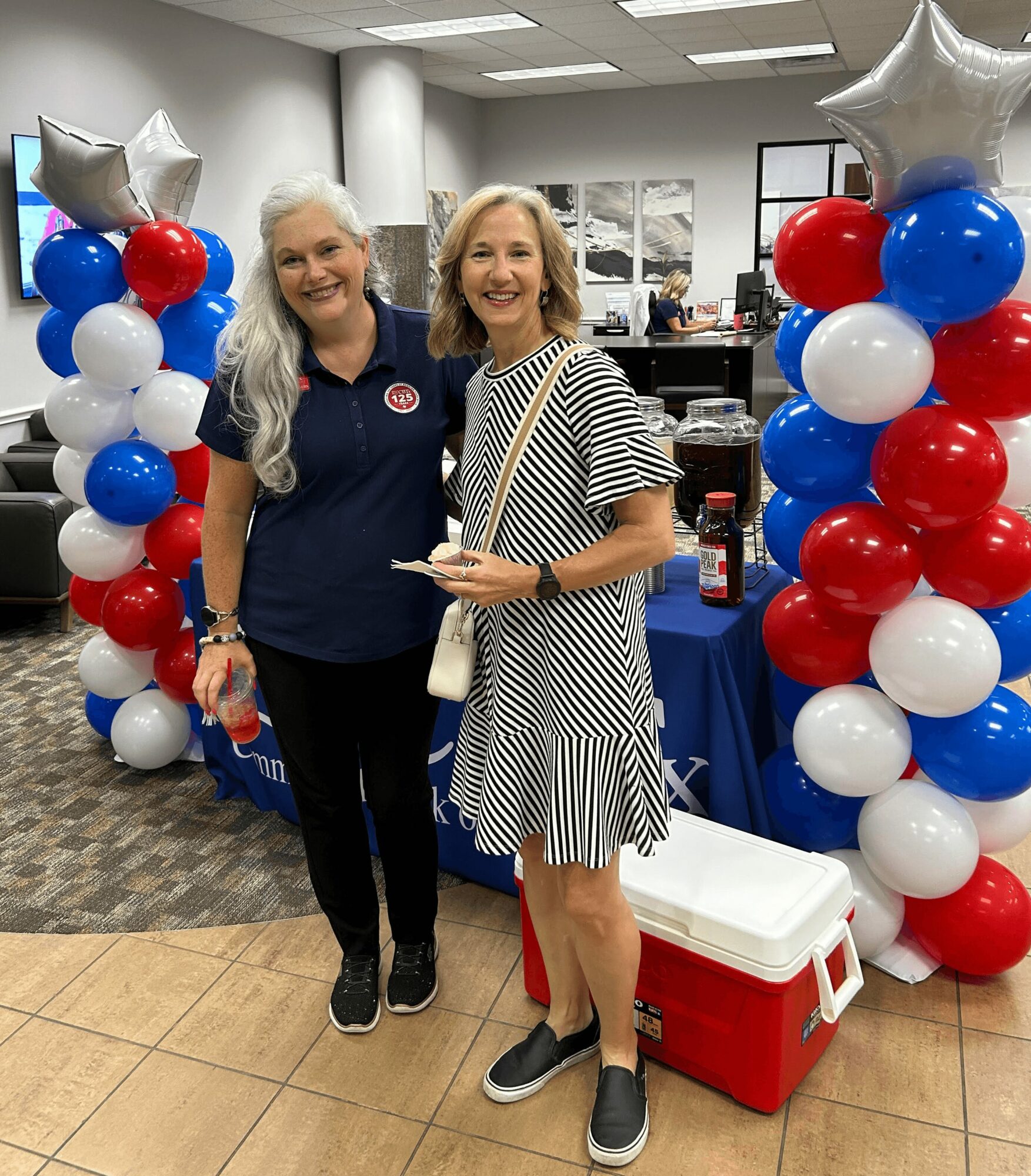 Two women smiling and standing between patriotic balloon columns with star-shaped balloons, in an indoor setting with tables and windows.