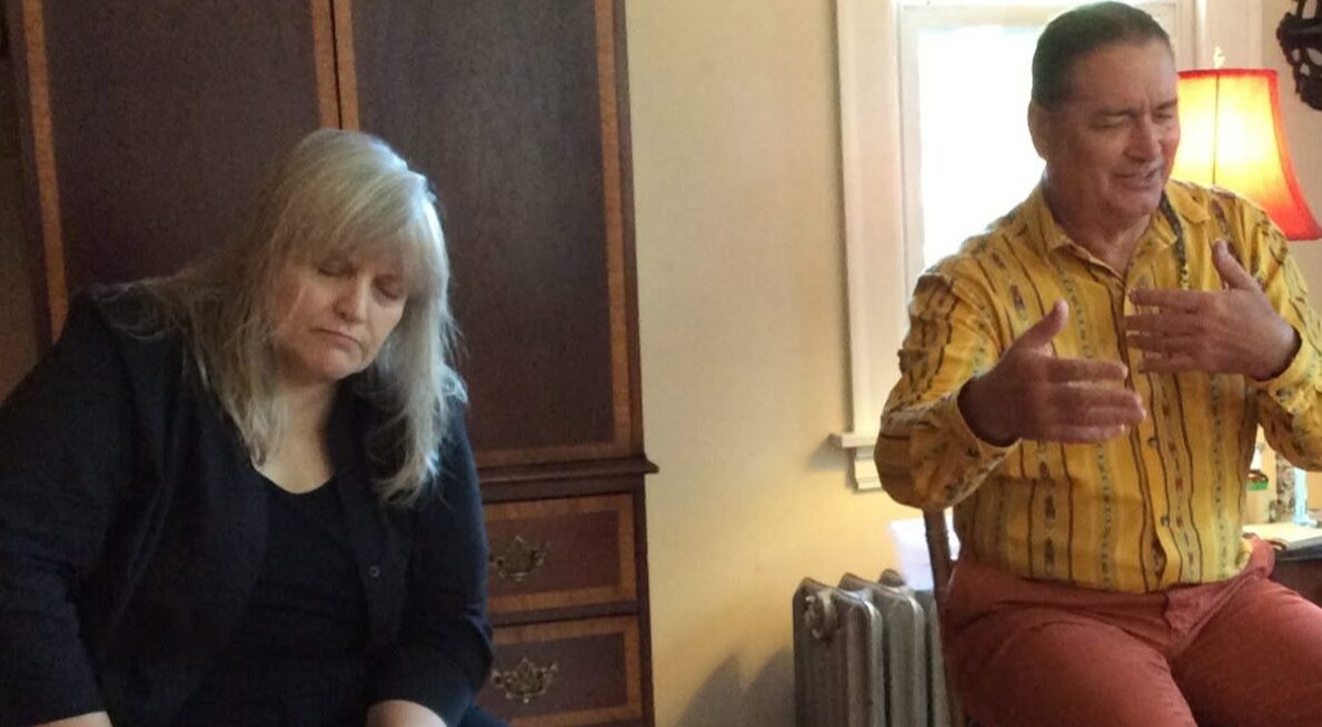 Two people sitting indoors, one woman with long blonde hair and a man in a yellow shirt, smiling and gesturing.
