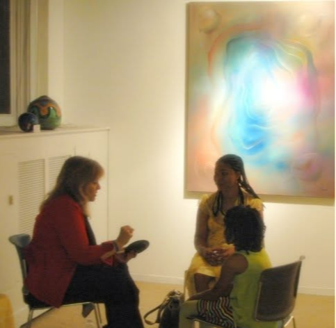 Two women sit and converse in an art gallery with a colorful abstract painting behind them.