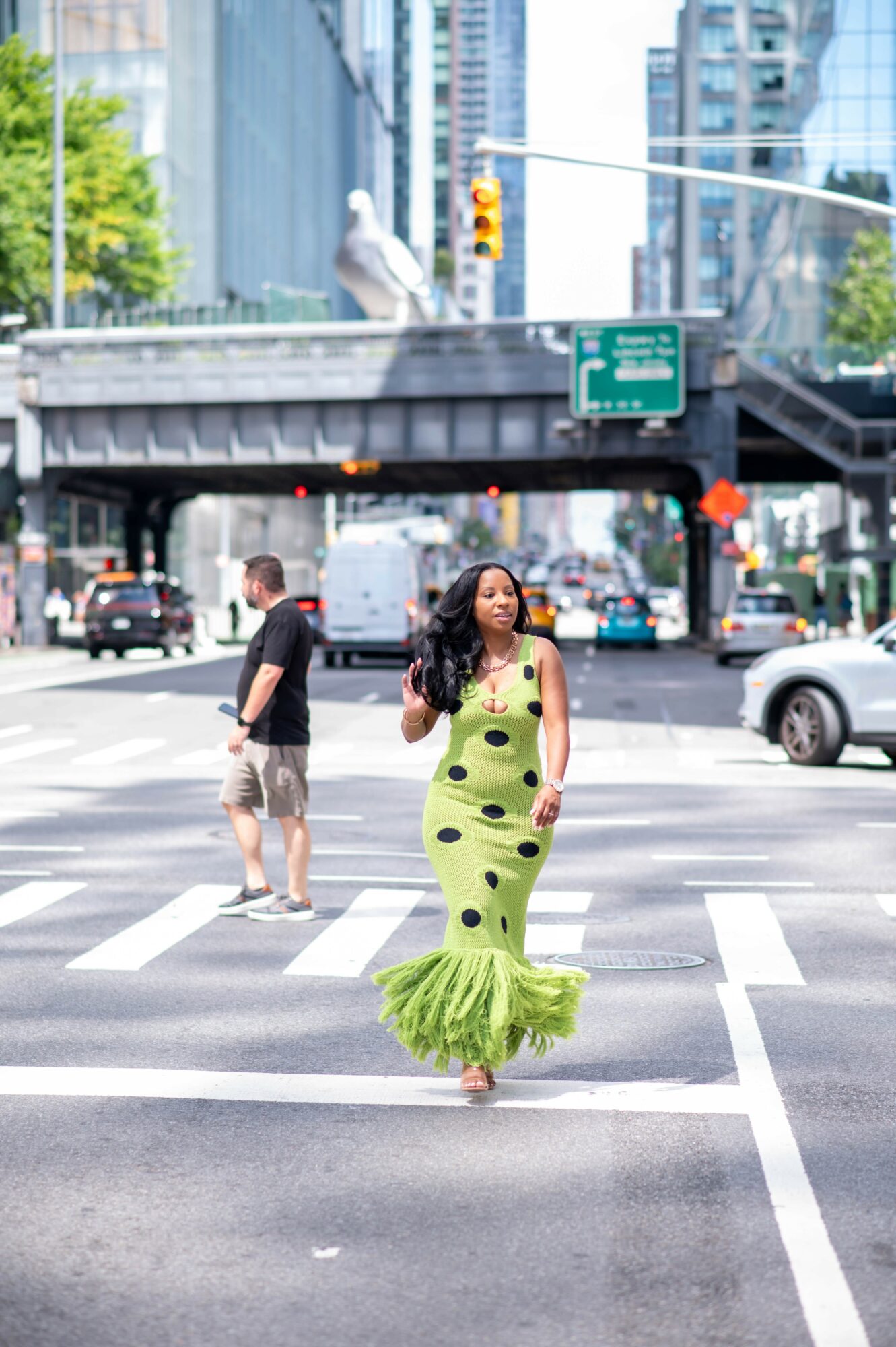 Woman in green polka dot dress walking across city street with cars and buildings in background.