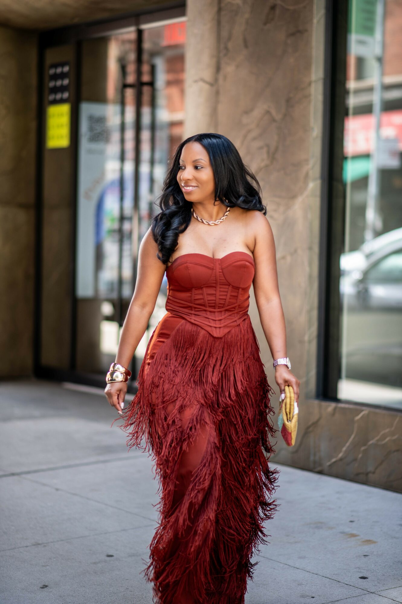 Woman in red strapless dress with feathered skirt walking outside, smiling, holding a yellow clutch, in front of glass building