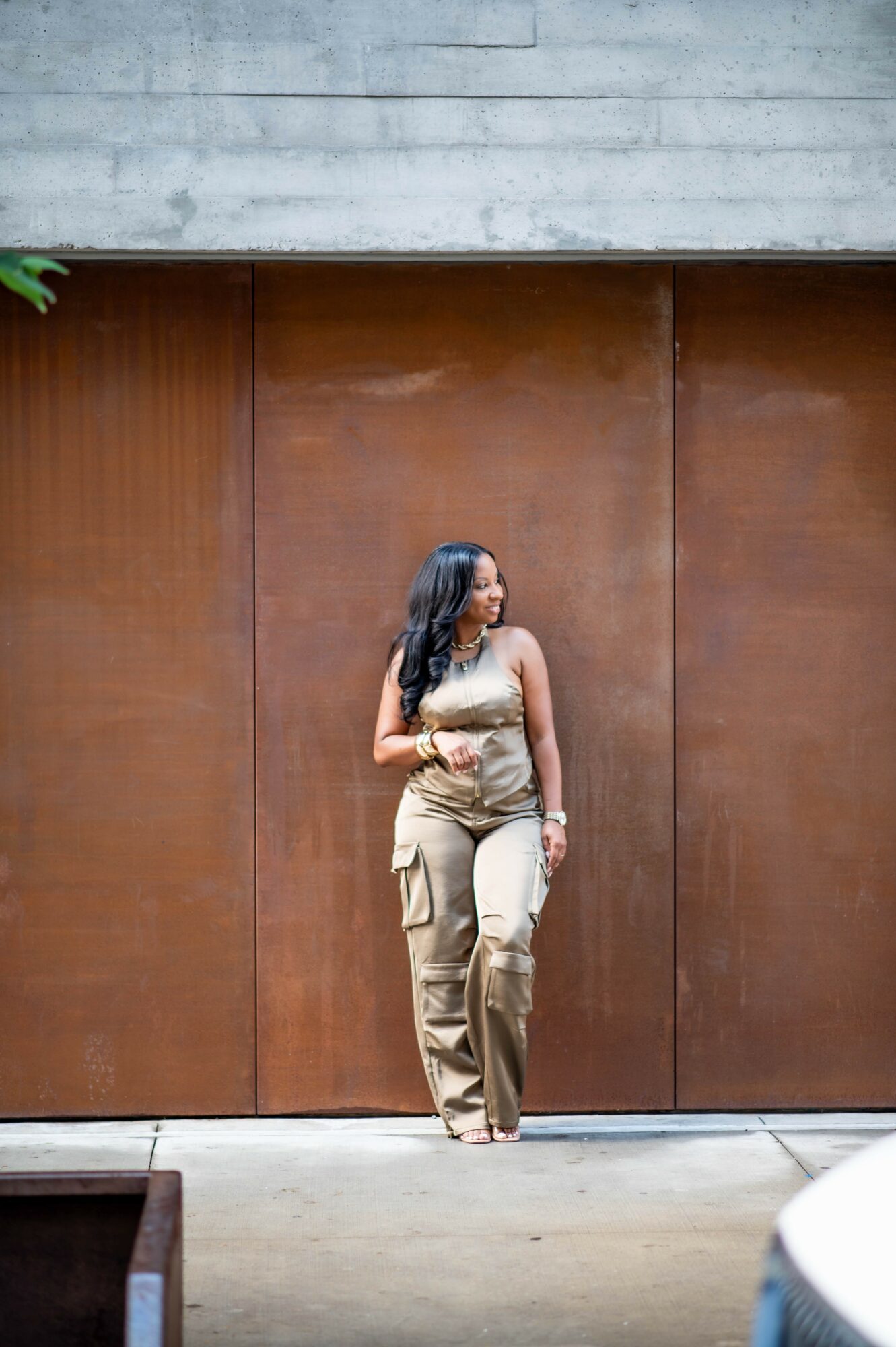 Woman standing in front of a large brown wooden door, looking to her right, wearing beige cargo pants and a sleeveless top.