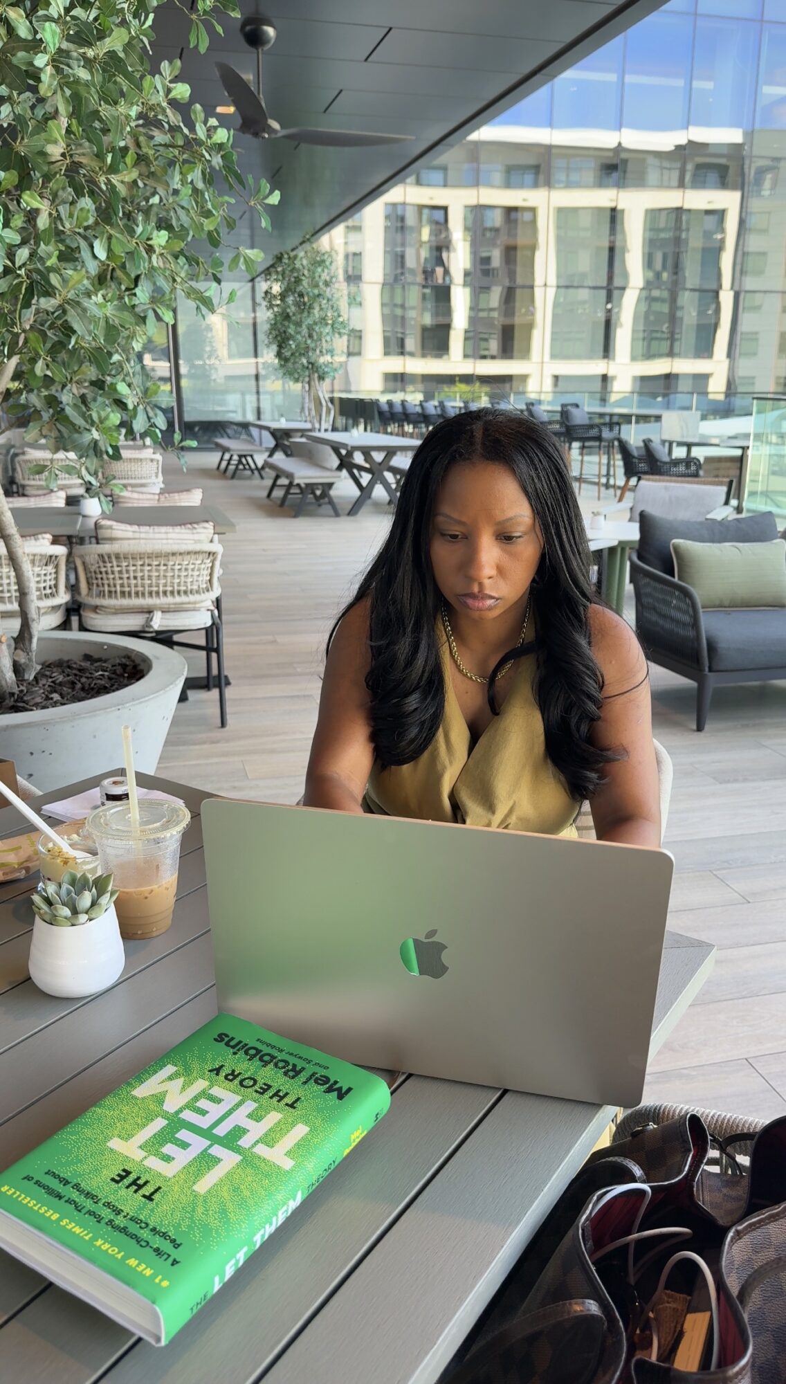 Woman sitting at a table with a laptop, a green book, and a drink in a modern indoor space with large windows.