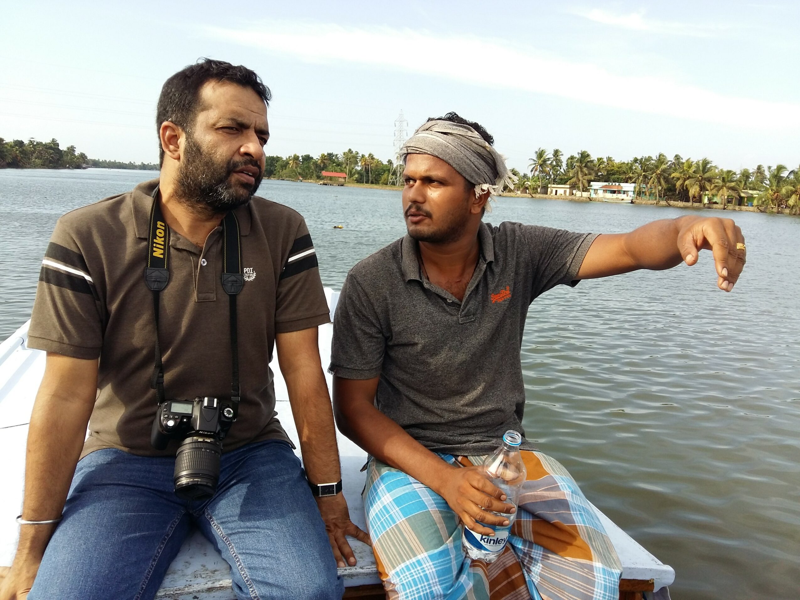 Two men sit by a body of water, one with a camera around his neck, the other holding a drink, with trees in background.