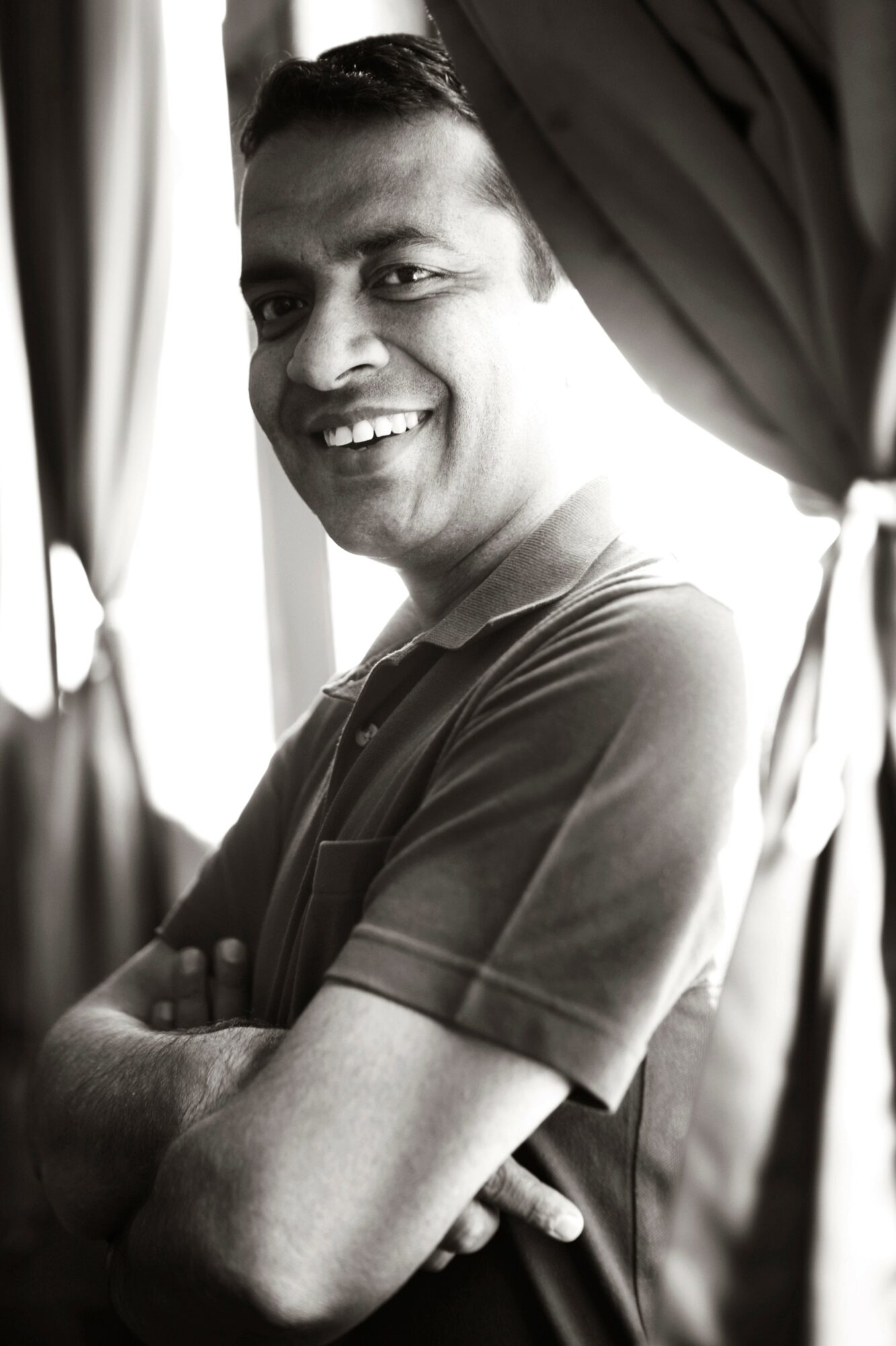 Smiling man with arms crossed, standing near curtains, in black and white.