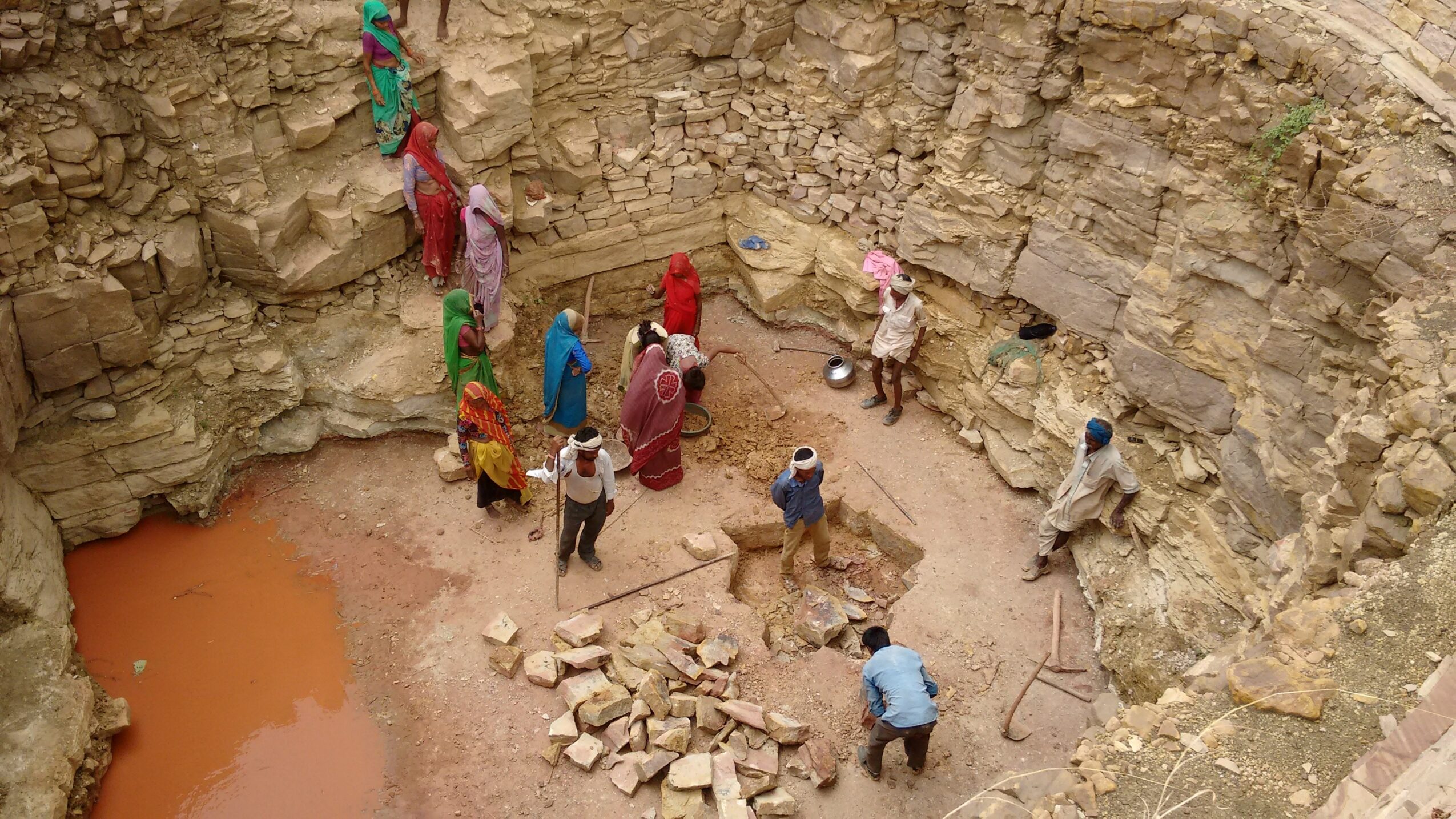 Group of people inside a circular excavation site with rocky walls, some sitting and others standing, with a small pool of water.